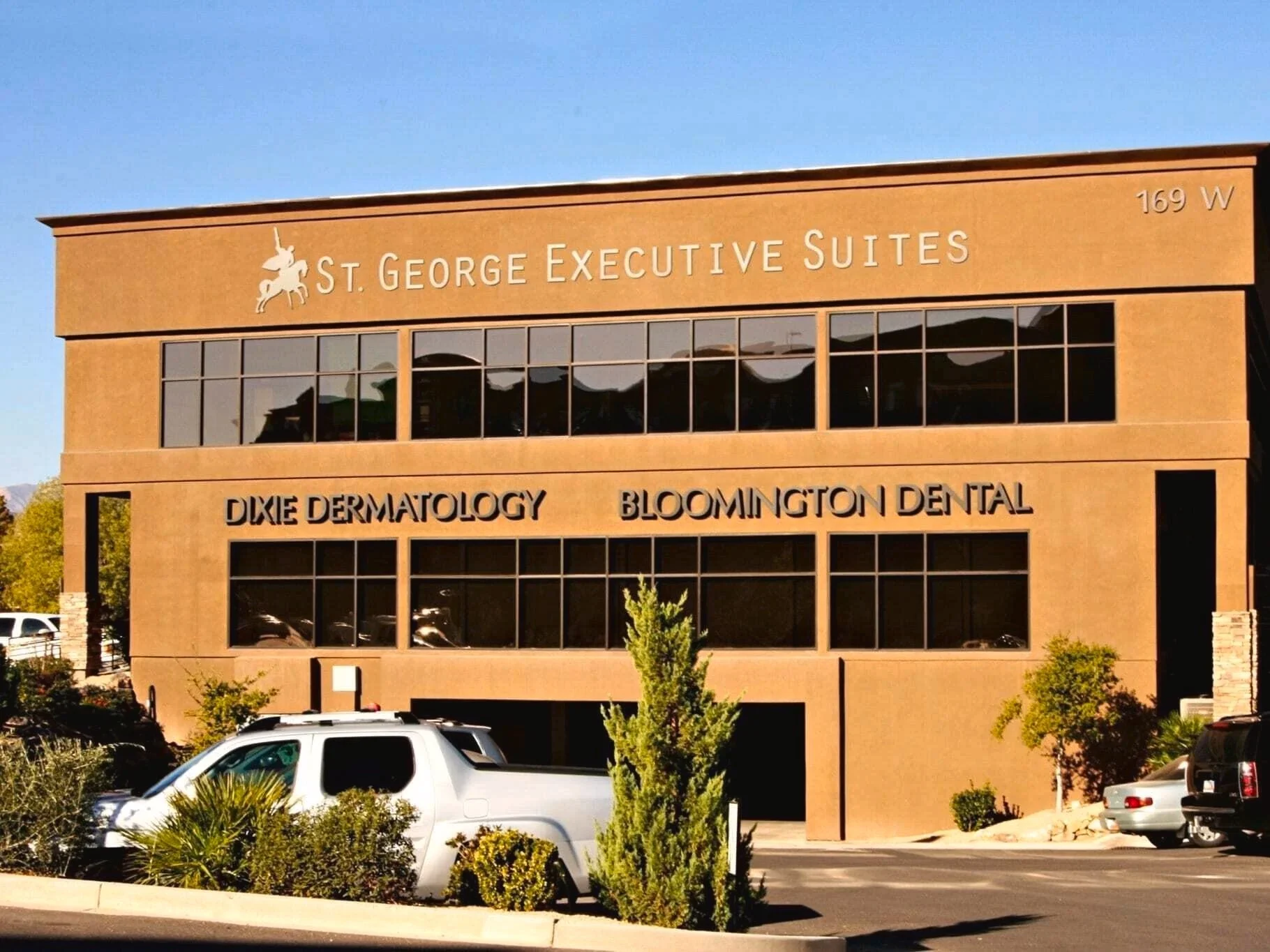 A three-story commercial building with a sign reading 'St. George Executive Suites' on top and businesses 'Dixie Dermatology' and 'Bloomington Dental' on the second floor. There are several cars parked in front of the building along with some small bushes and trees.