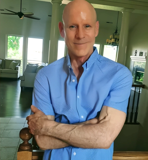 A bald man with light skin and a slight smile, wearing a blue short-sleeved shirt, standing in a bright, spacious living room with large windows and white furniture.