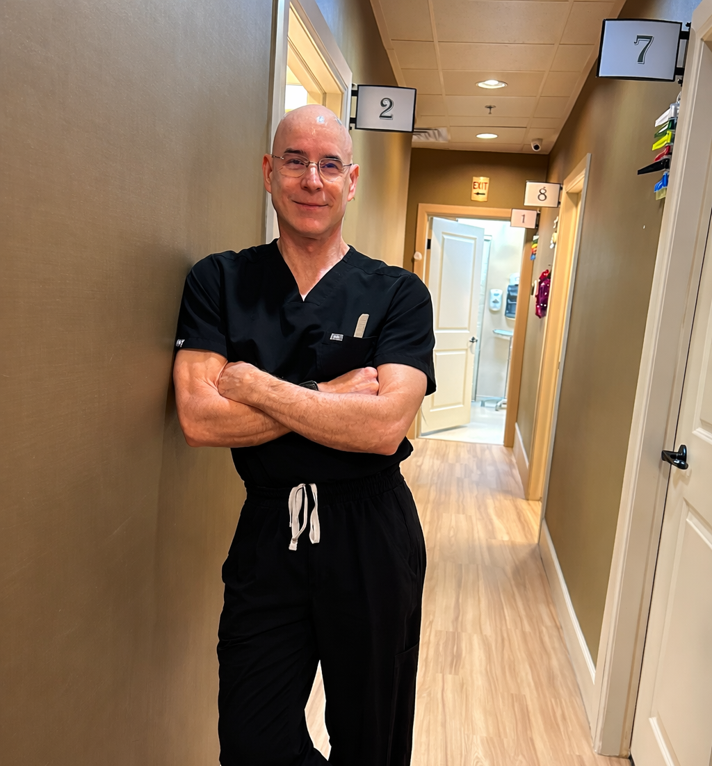 A man in a black medical uniform standing in a hallway of a healthcare facility with arms crossed, smiling.