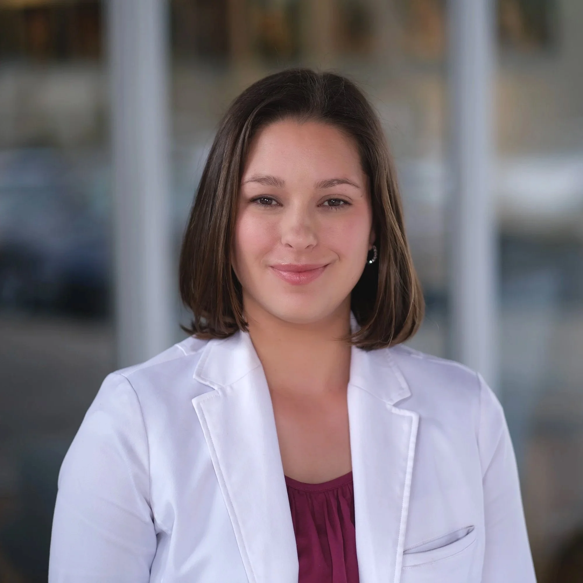Portrait of a woman with shoulder-length brown hair, wearing a white blazer and a maroon top, smiling gently in an outdoor setting with blurred background.