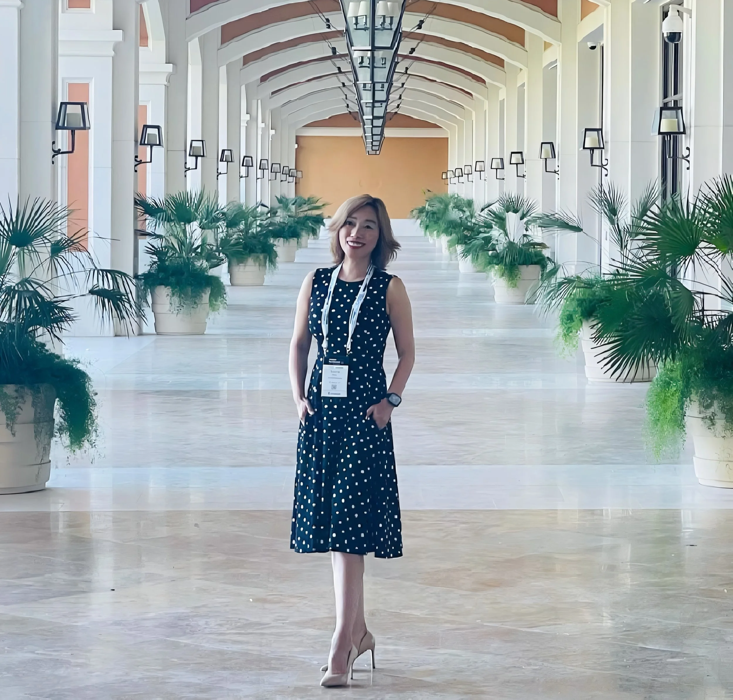 A woman in a navy blue polka dot dress and beige high heels standing in a spacious, well-lit corridor with arched ceiling, wall-mounted lights, and potted plants lining the sides.