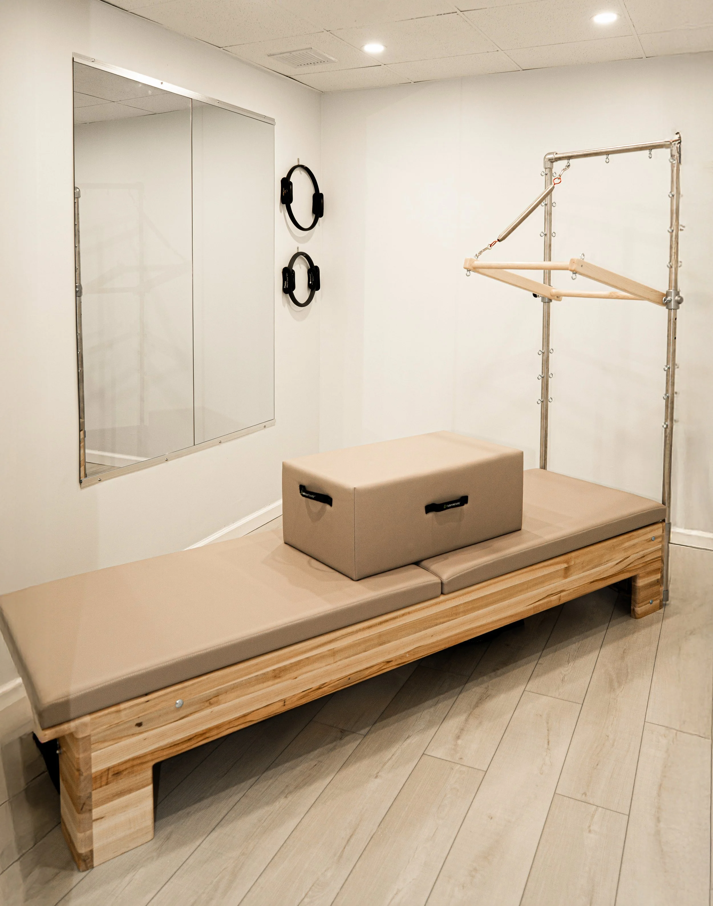 A beige padded bench with a workout box on top, exercise equipment including a padded workout frame with bars and pulleys, a mirror, and two black headphones hanging on the wall in a small gym or workout room.