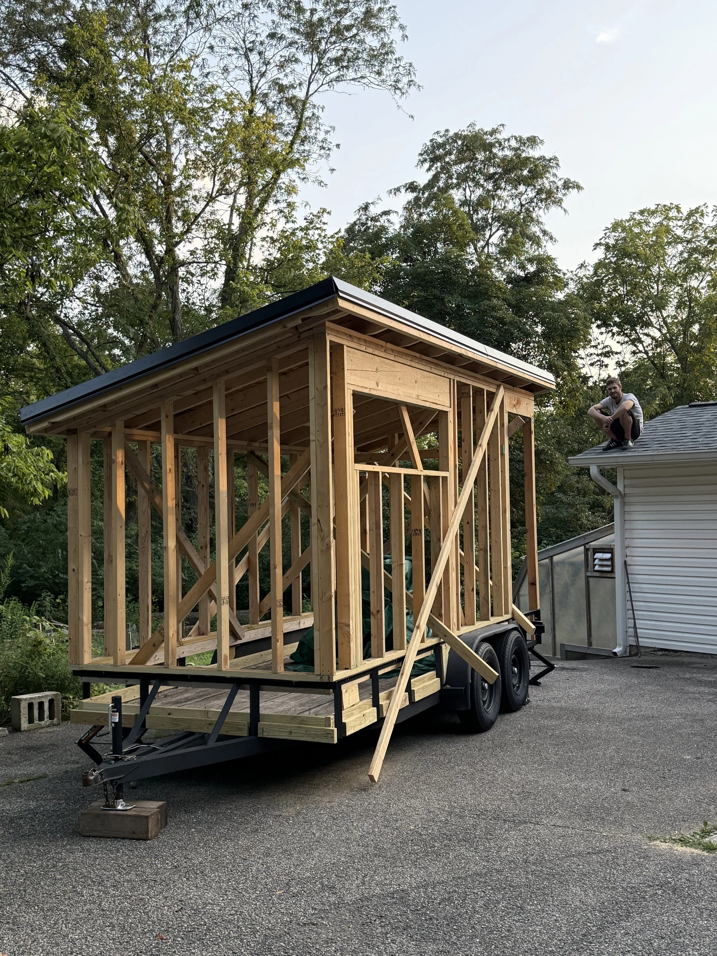 A mobile sauna under construction with a wooden frame on a trailer, with a man sitting on the roof of an already finished house nearby, surrounded by trees.