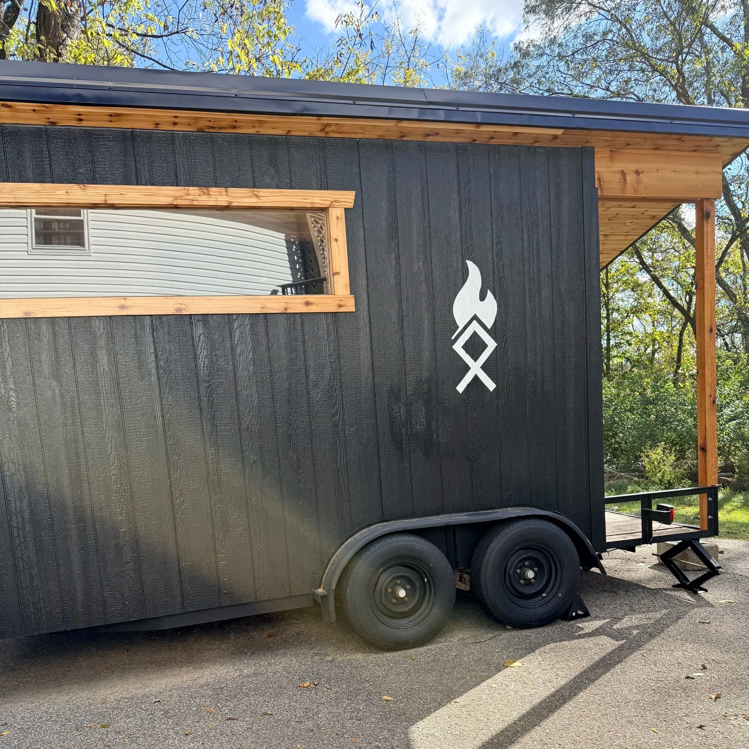 Black and cedar mobile sauna with a custom rune that looks like a white flame icon on the side, featuring a small window with wooden trim, double black wheels, and a rear step, parked on a paved surface surrounded by trees and a blue sky.