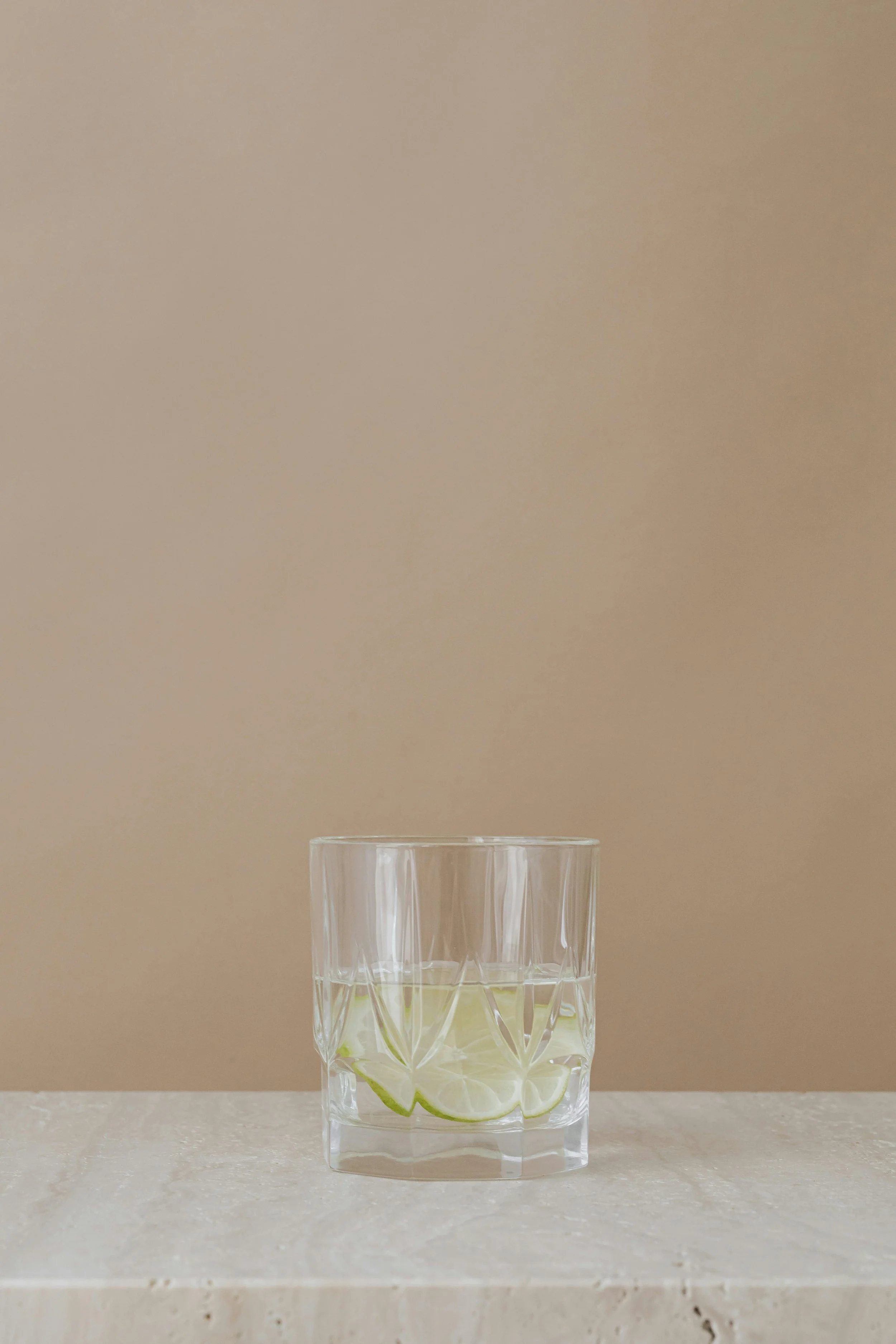 A clear glass tumbler with a faceted design, filled with water and slices of lime, sitting on a light-colored surface against a beige wall.