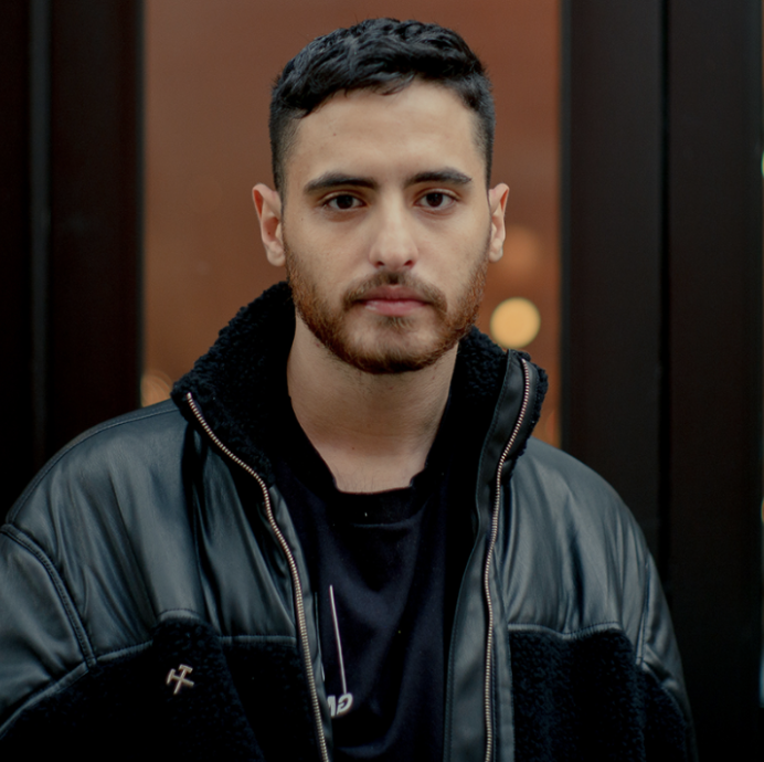 A portrait of a young man with short dark hair, wearing a black collared shirt, smiling against a dark background.