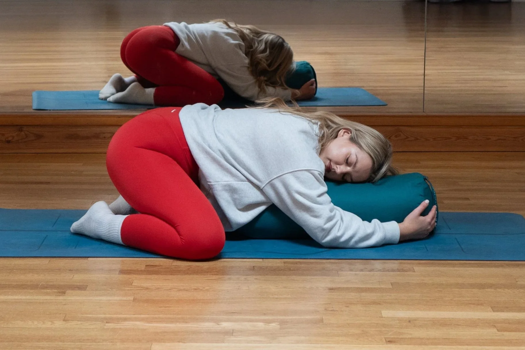 A woman is practicing yoga on blue mats in front of a mirror. They are in child's pose, resting with their foreheads on a yoga bolster, eyes closed, and hands clasped on the mat.