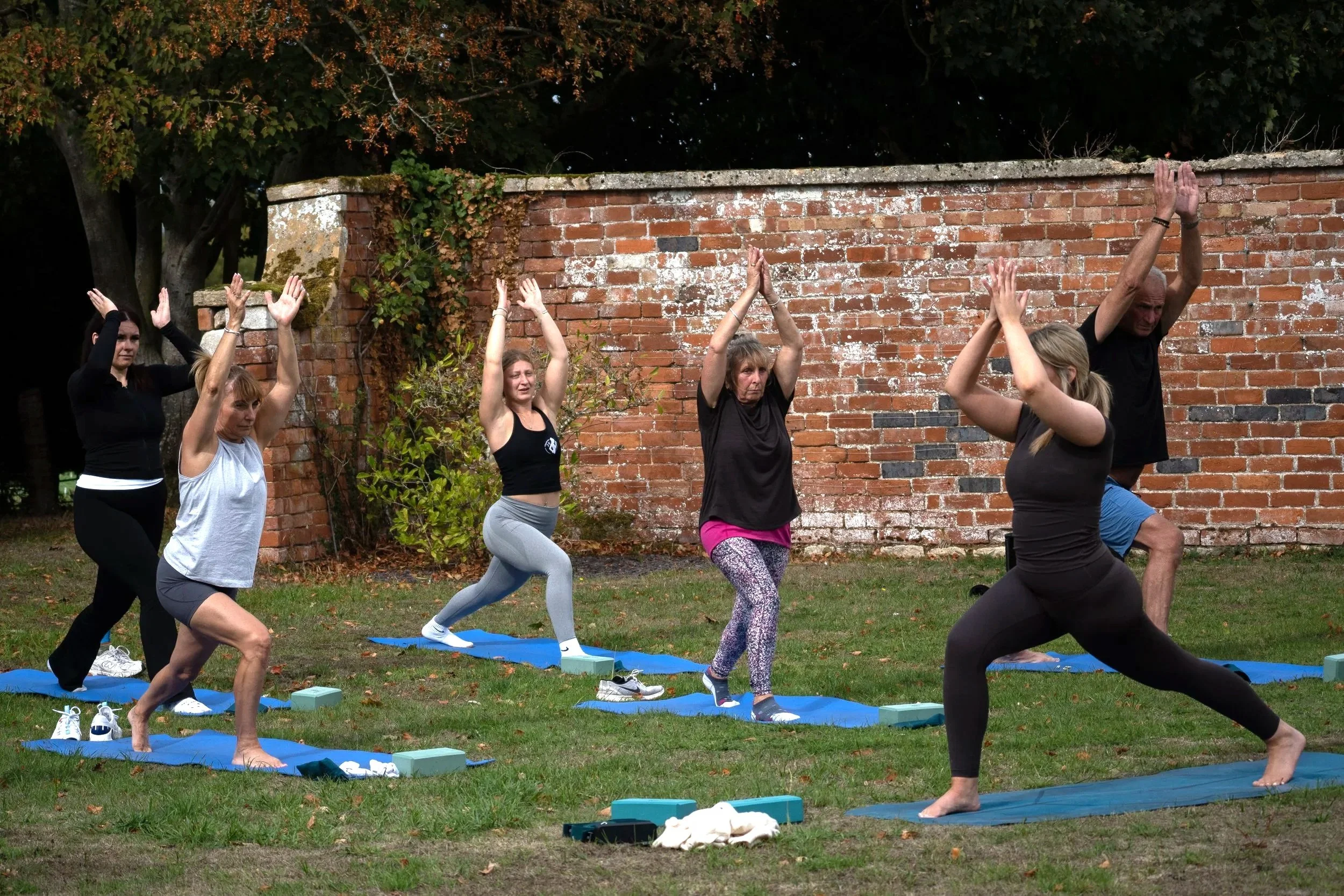 Group of six people practicing yoga outdoors on blue mats, with a brick wall and trees in the background.