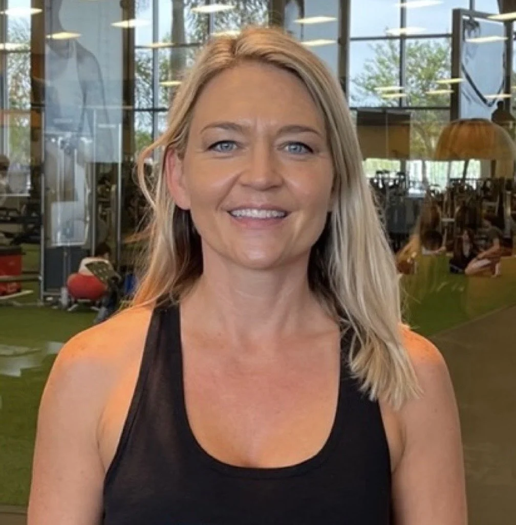 A woman with blonde hair and blue eyes smiling at the camera inside a gym with large windows.