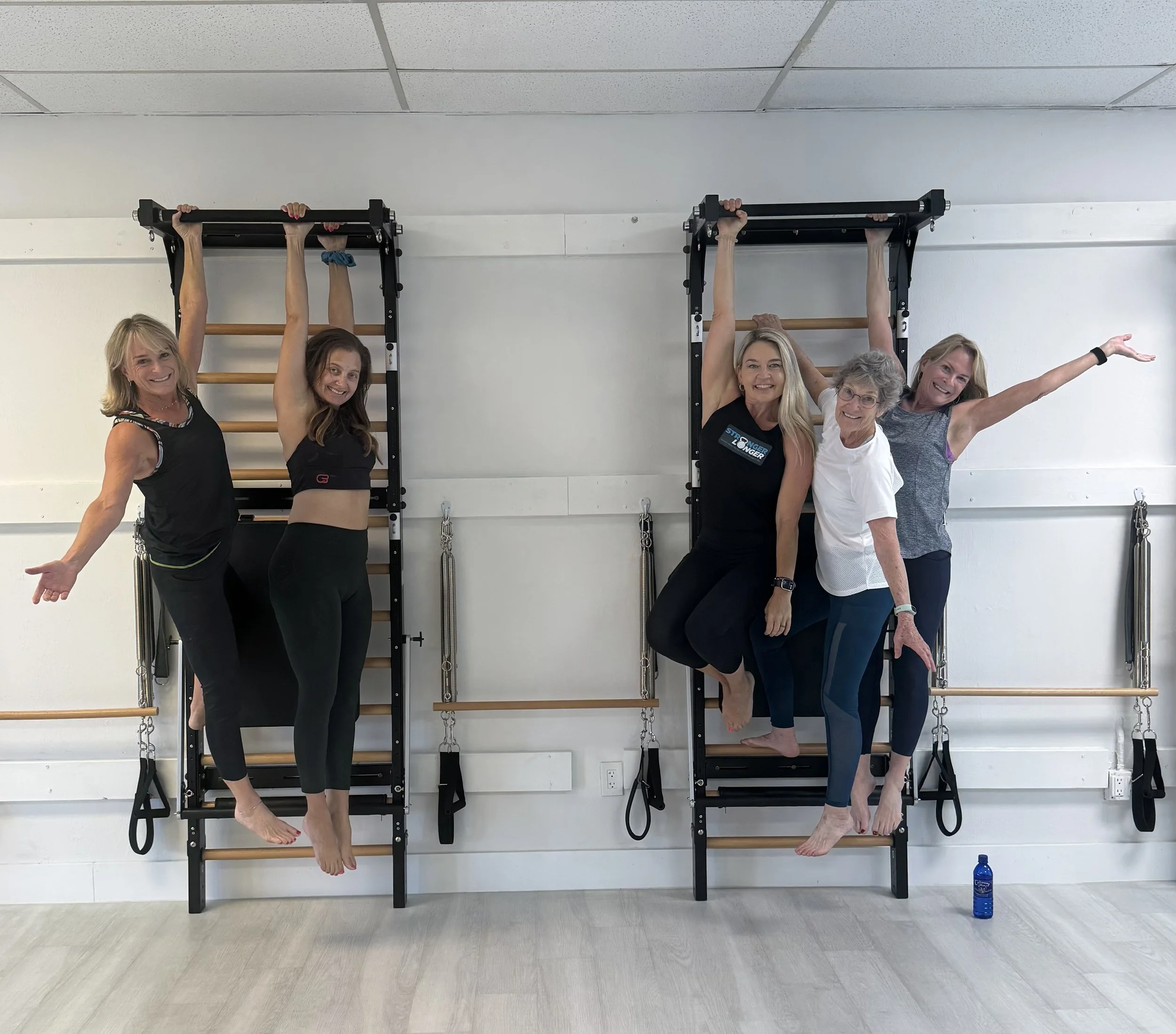 Group of five women smiling and posing with their arms raised and hanging from gym wall bars in a fitness studio, with a water bottle on the floor.