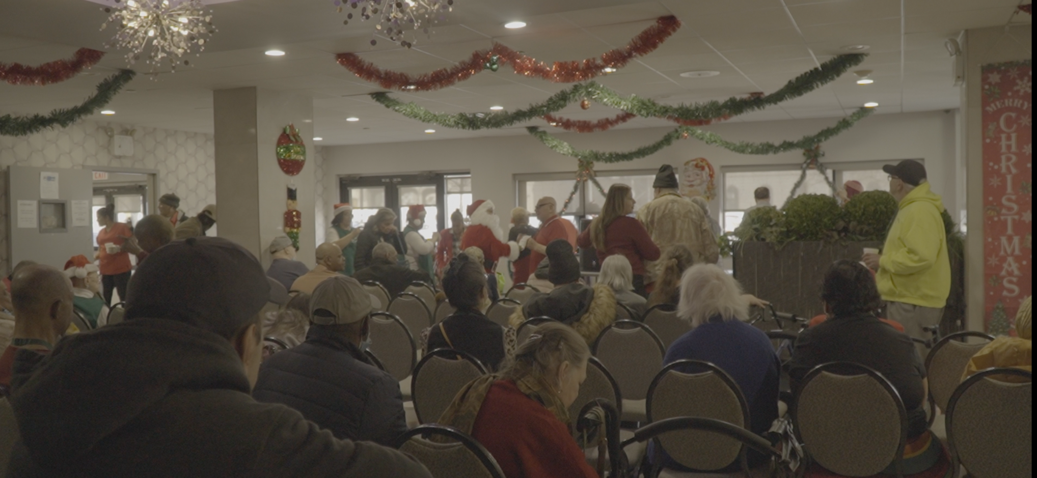 Indoor Christmas gathering with people in festive attire, decorations hanging from the ceiling, including garlands and a star-shaped light fixture, and holiday-themed wall decor.