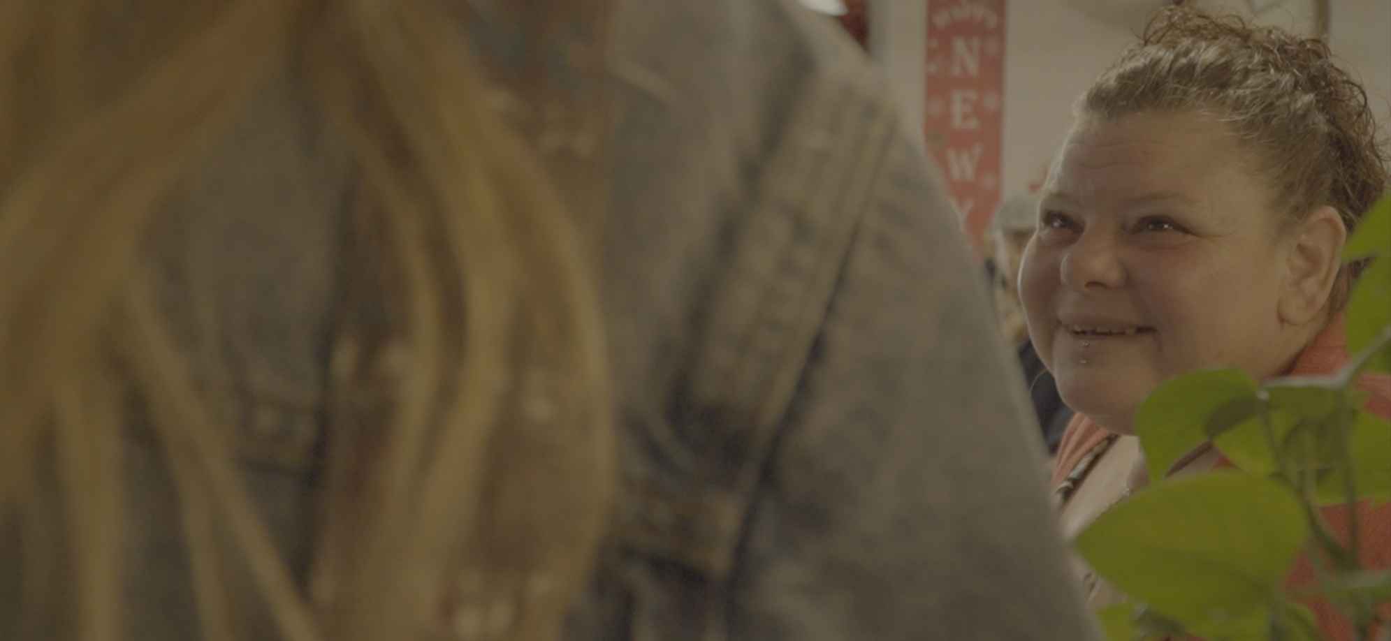 A woman with curly hair and a lip piercing smiling and talking to a person with blonde hair, both in an indoor setting with plants and decorations.