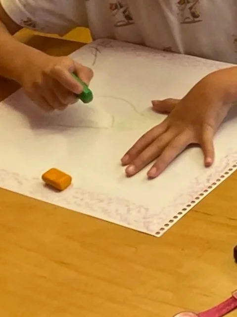 Person drawing or coloring on a large sheet of paper with a green crayon, with another orange crayon on the table.