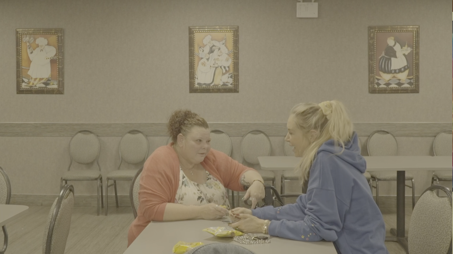 Two women sitting at a table in a room, with the woman on the left handing something to the woman on the right.