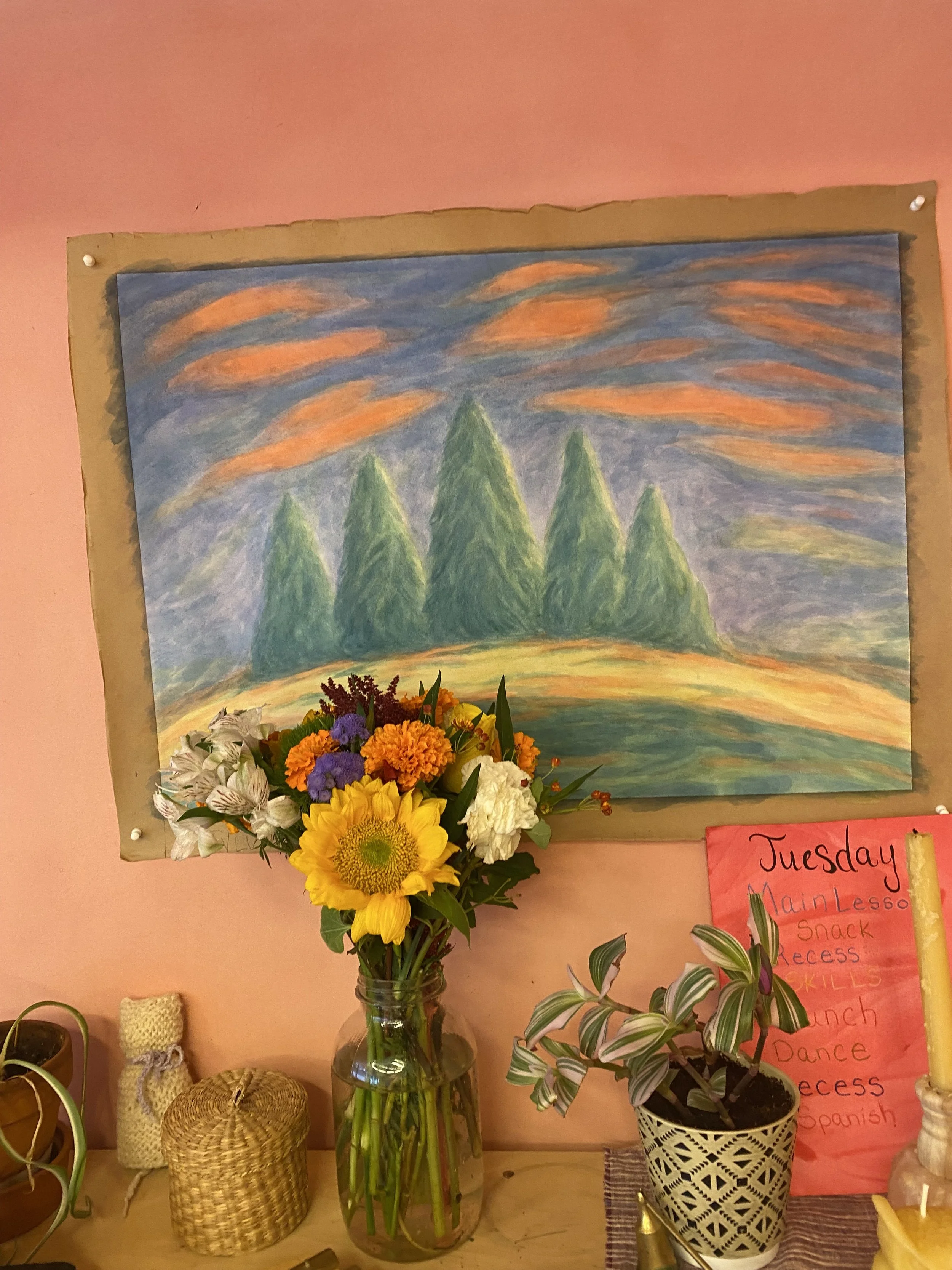 A colorful indoor scene featuring a painting of a landscape with trees and a sky, a floral arrangement in a clear glass vase, a potted plant with variegated leaves, and a pink note with a daily schedule and a yellow candle.