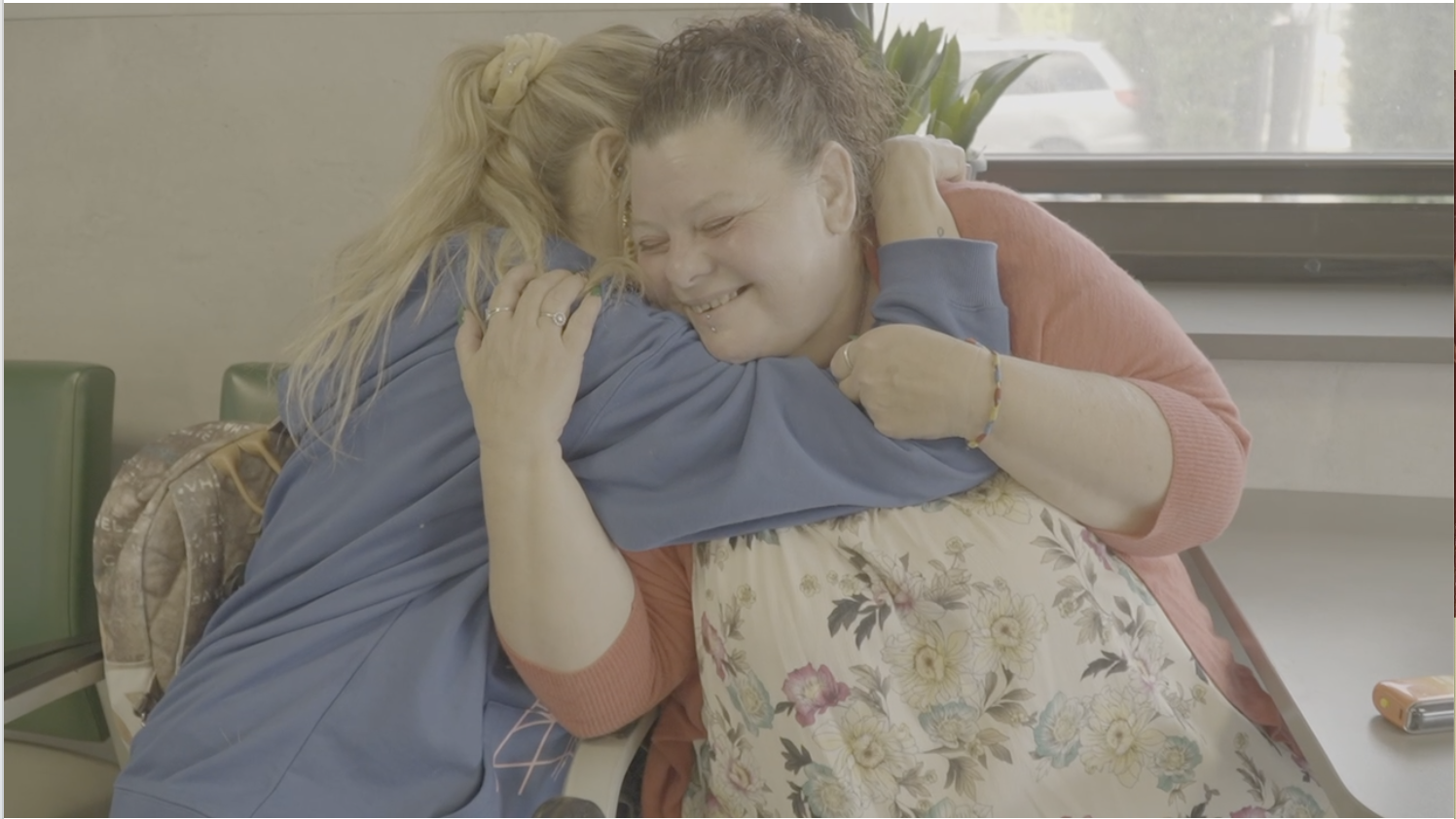 Two women hugging and smiling, one with blonde hair and the other with curly red hair, inside a room with a window showing a car outside.