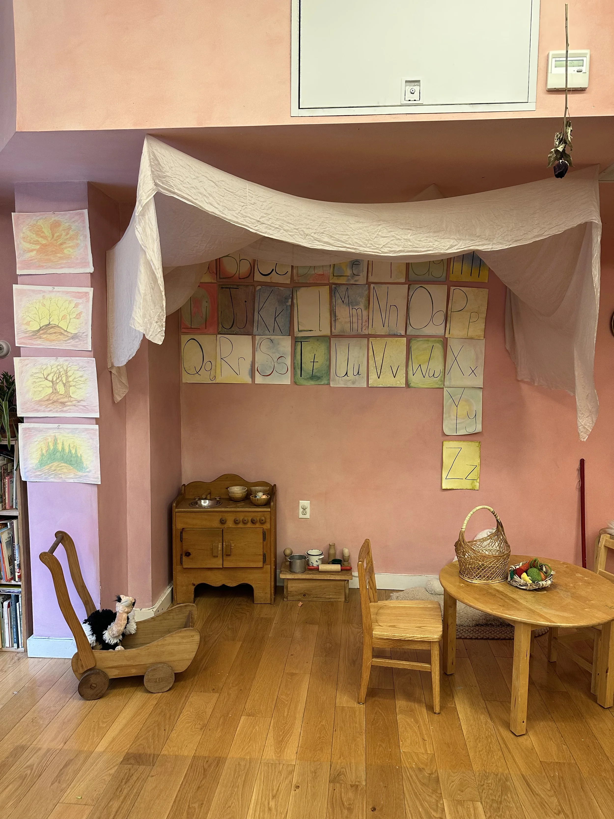 A cozy children's play area with pink walls, a small wooden table and chairs, a wooden toy cabinet, and child's artwork on the wall. There is a plush toy in a wooden cart and a basket with toys on the table, with paper and drawings hanging on the wal