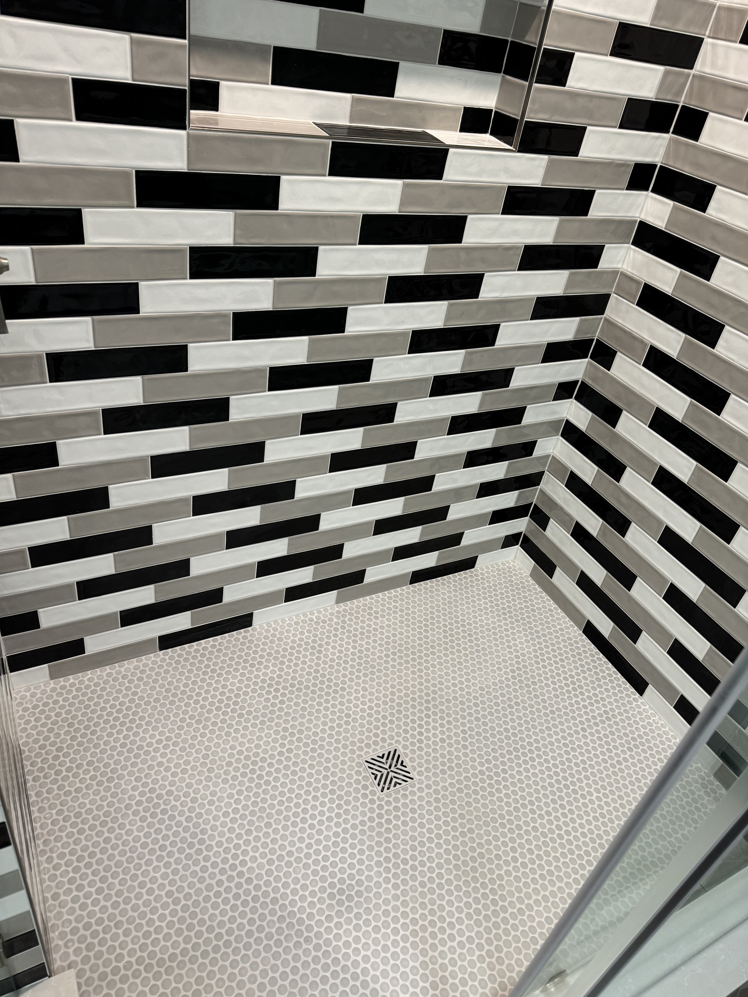 A corner of a bathroom shower with black, white, and gray rectangular tile walls and a white hexagonal tile floor with a square drain in the center.