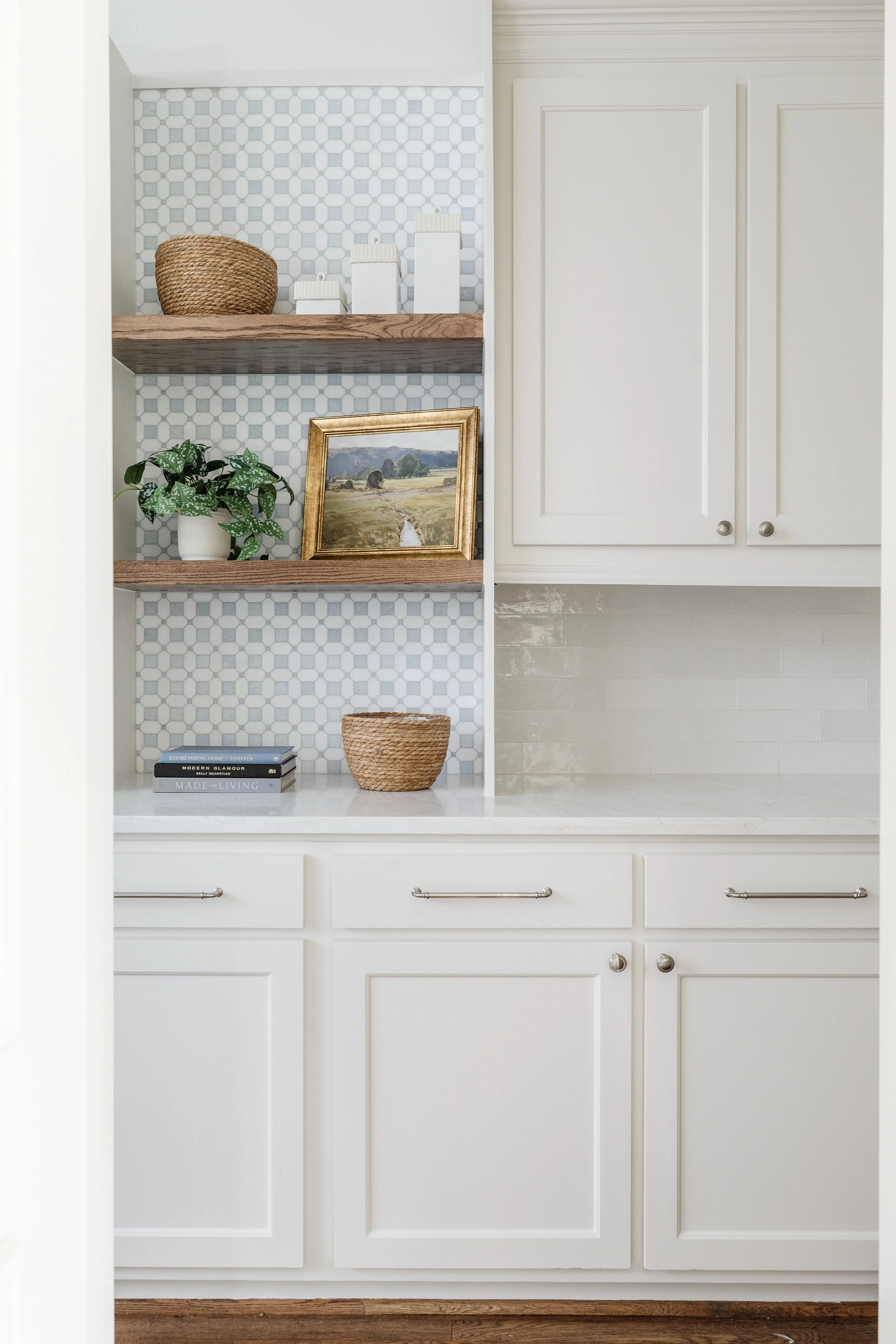 Kitchen with white cabinets, open wooden shelves with baskets, a framed landscape painting, potted plant, and books, all against a hexagon-tile backsplash.