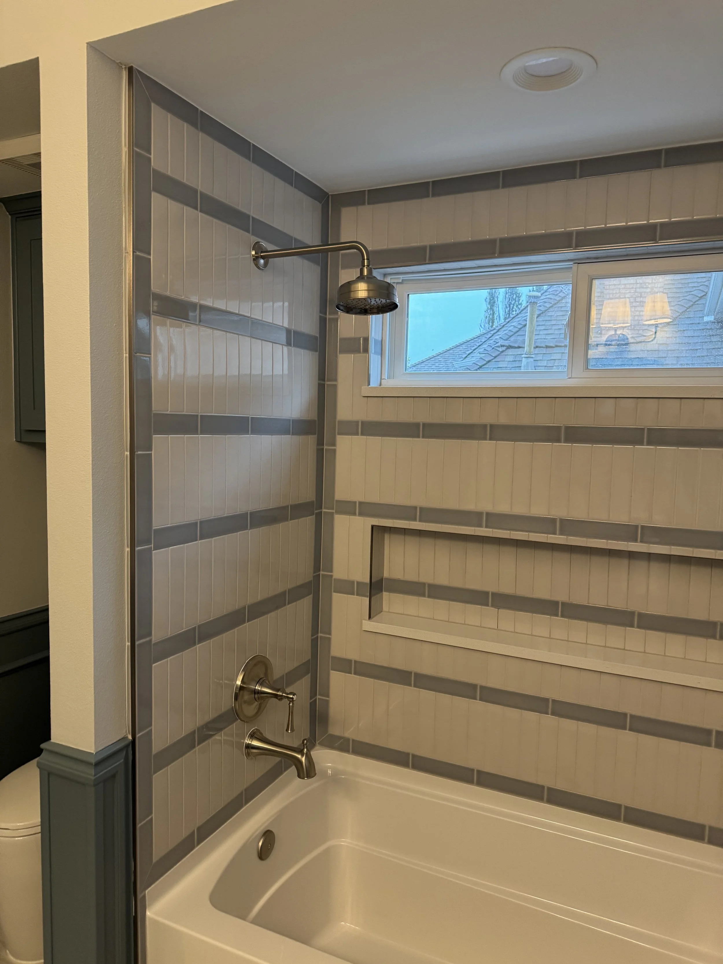 A bathroom with a bathtub, gray and white tile walls, a showerhead, and a small horizontal window showing a roof and two lamps outside.