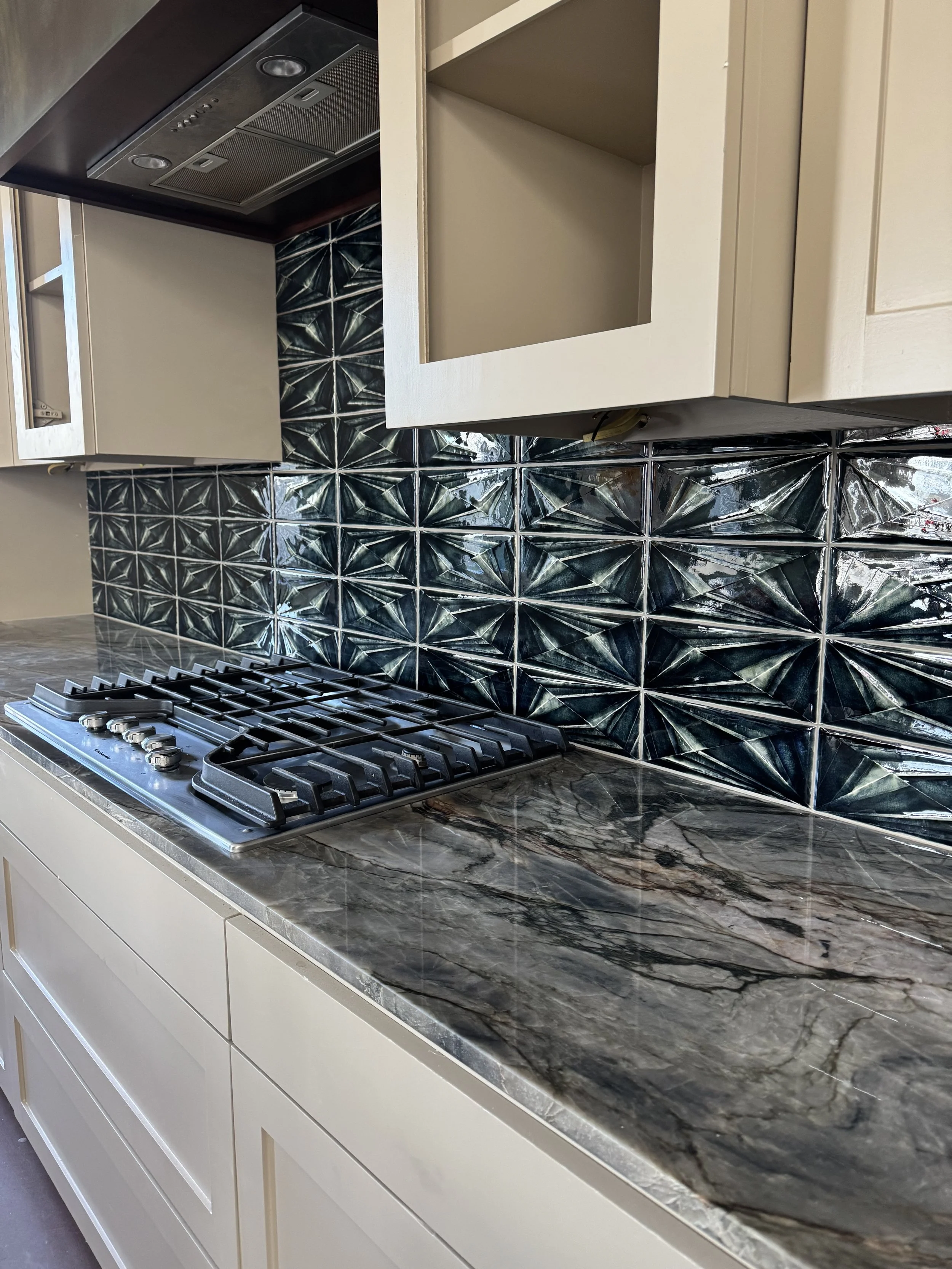 Kitchen countertop with a gas stove, beige cabinetry, black and silver geometric tile backsplash, and a marble pattern on the counter.
