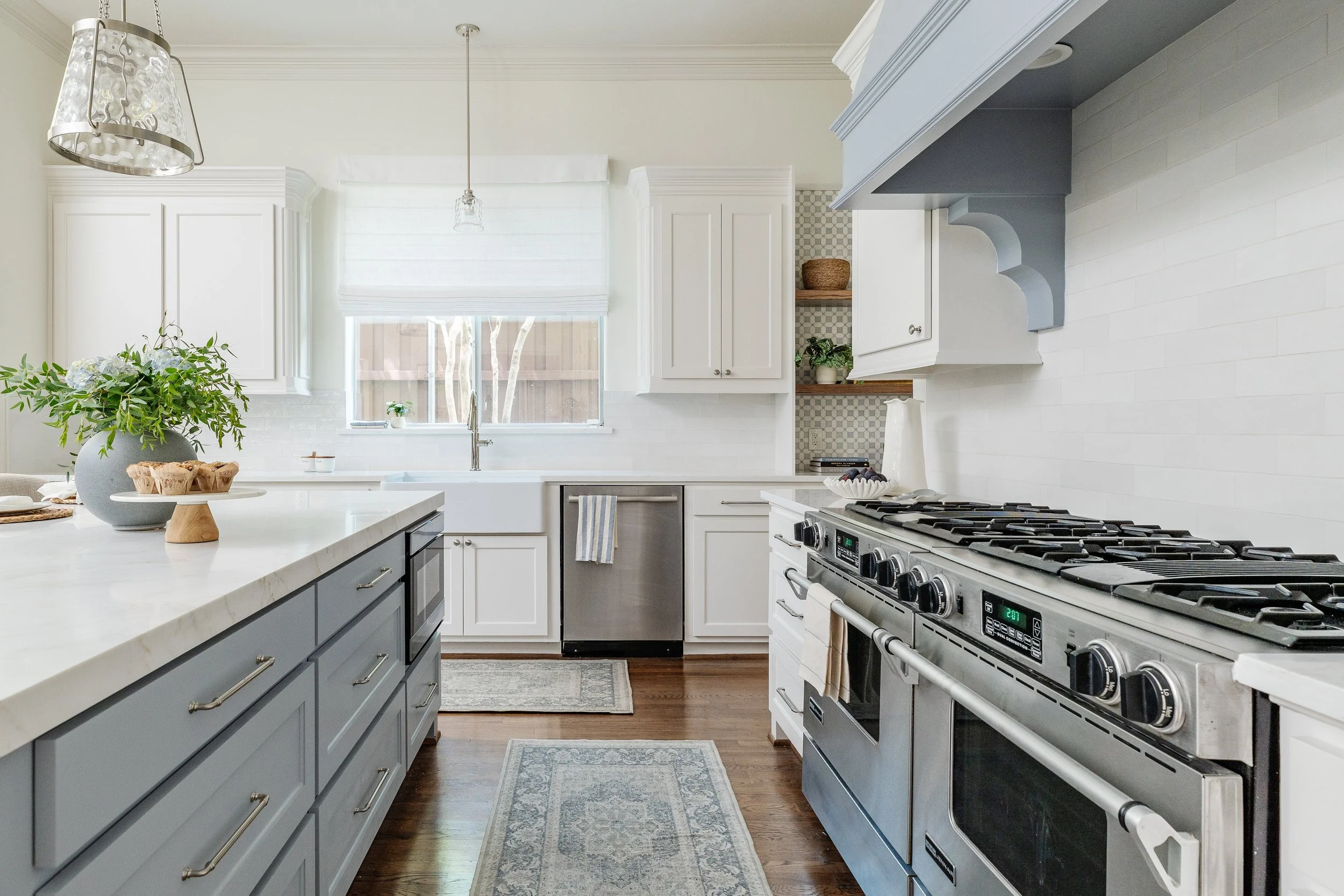 A bright, modern kitchen with white cabinets, a large stainless steel stove, a white marble countertop, a window above the sink, and decorative elements like a large vase with green foliage, a cake stand, and a rug on a dark wood floor.