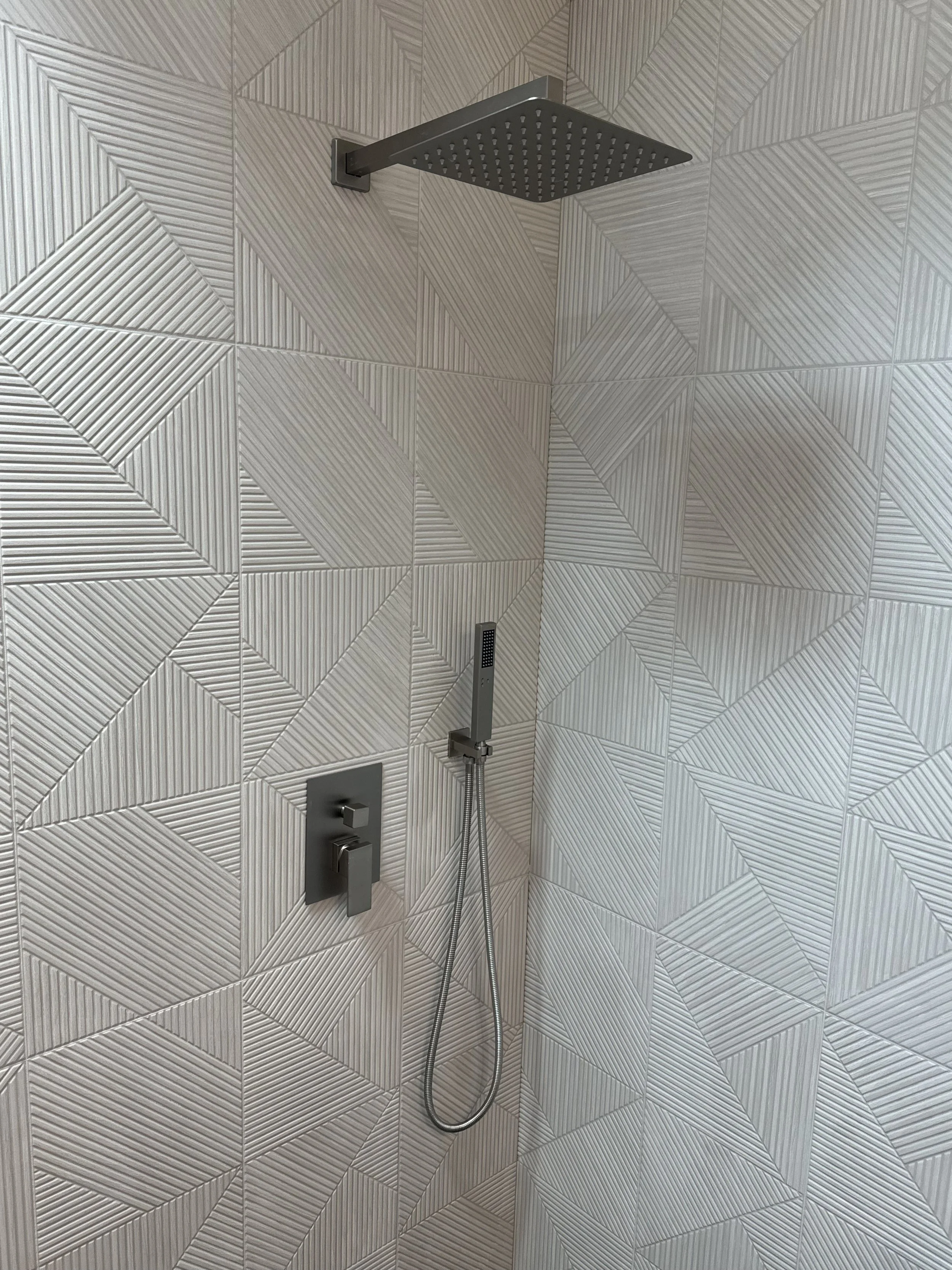 Modern shower with a square rain showerhead, a handheld showerhead, and a control panel, all installed on a tiled wall with geometric textured tiles.
