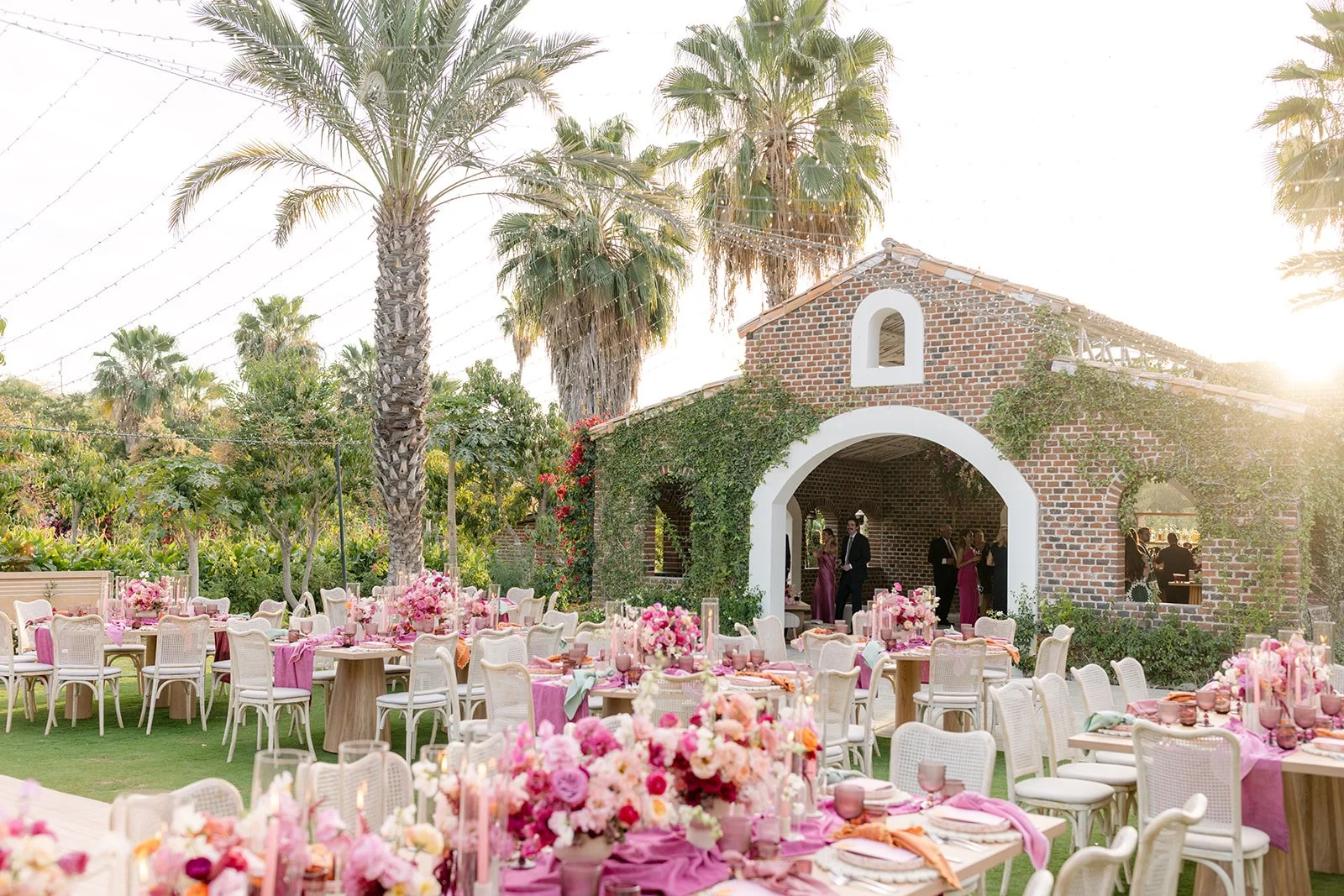 Outdoor wedding reception setup with round tables, pink floral centerpieces, and white chairs on a lush green lawn. In the background, a brick building with ivy and palm trees is visible, with guests mingling under a pergola.