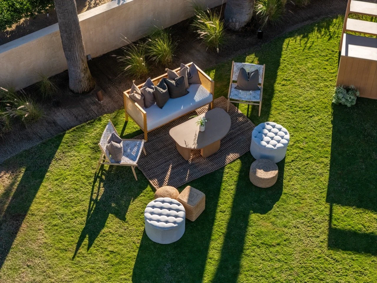 Aerial view of an outdoor backyard seating area with a white cushioned sofa with gray throw pillows, two matching armchairs, a rectangular coffee table, and additional small round ottomans on a grassy lawn.