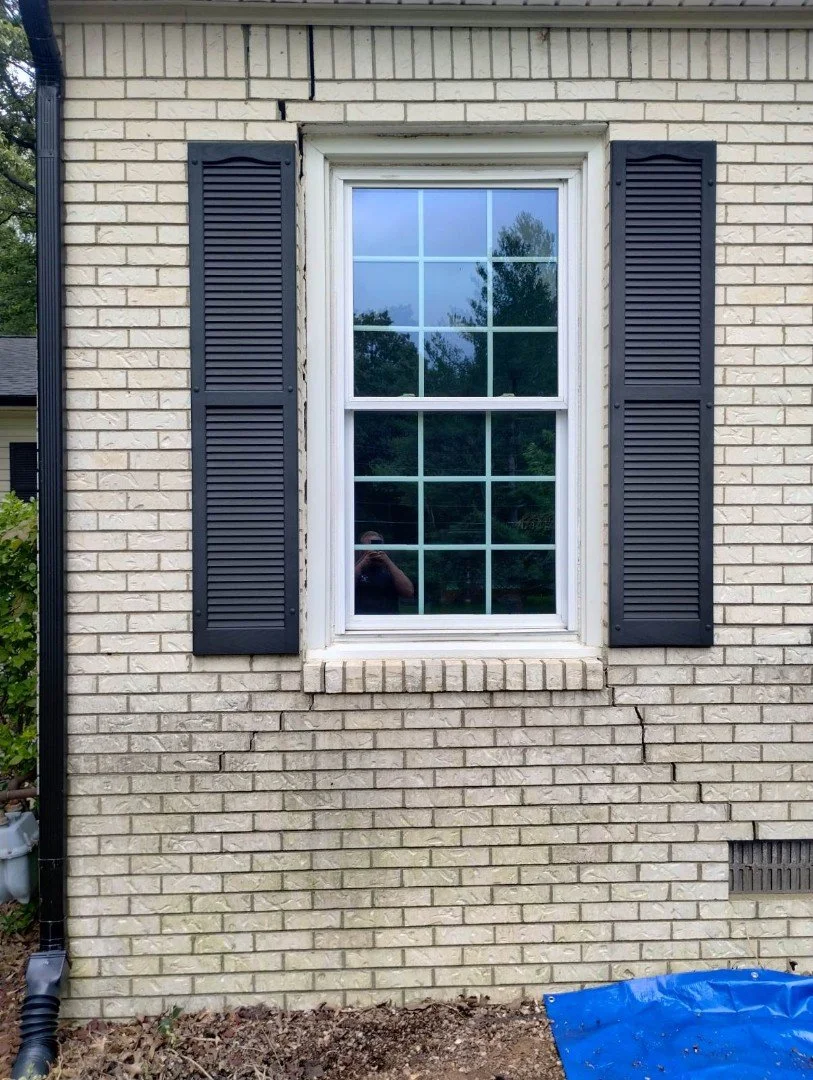 A window with white trim and black shutters on a brick house, reflecting trees and the person taking the photo.