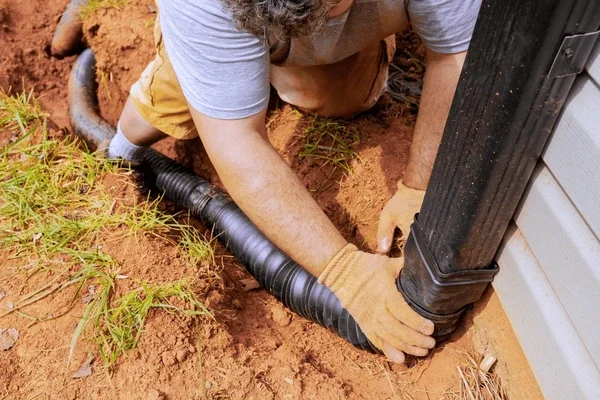 A person installing or repairing an underground drainage pipe next to a building foundation, using gloves to handle a black corrugated pipe.