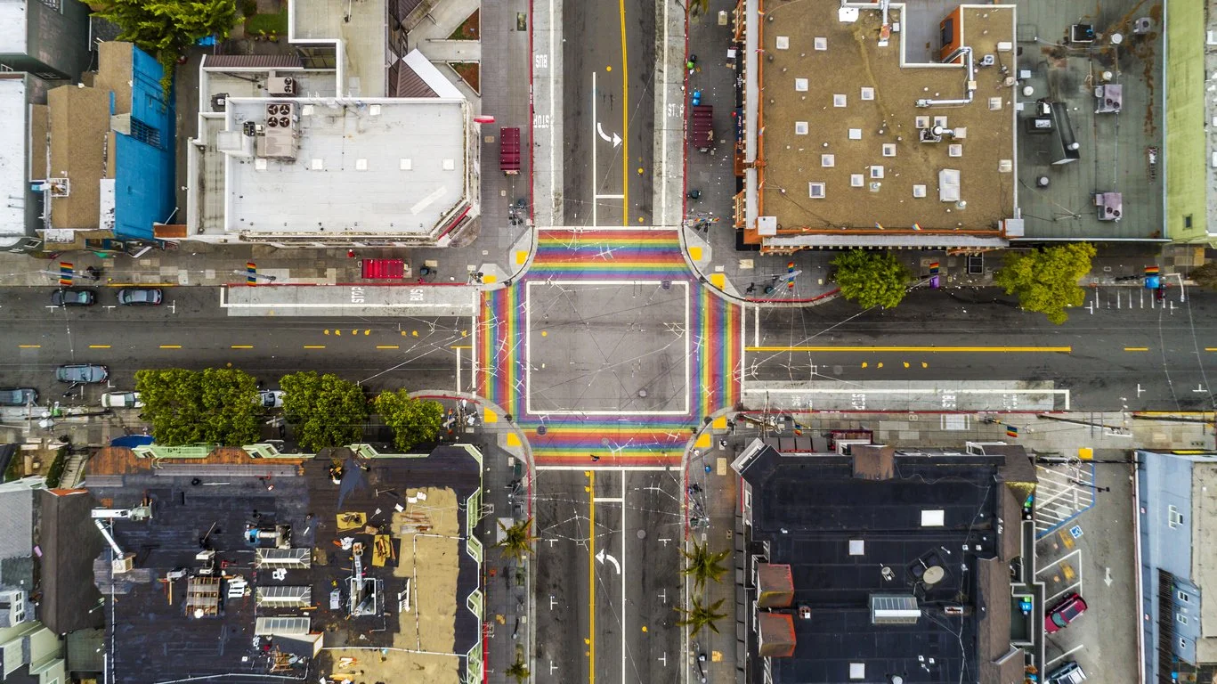 An aerial view of a city intersection with four intersecting rainbow-colored crosswalks as well as surrounding buildings, trees, and parked cars in the Castro neighborhood of San Francisco, California.
