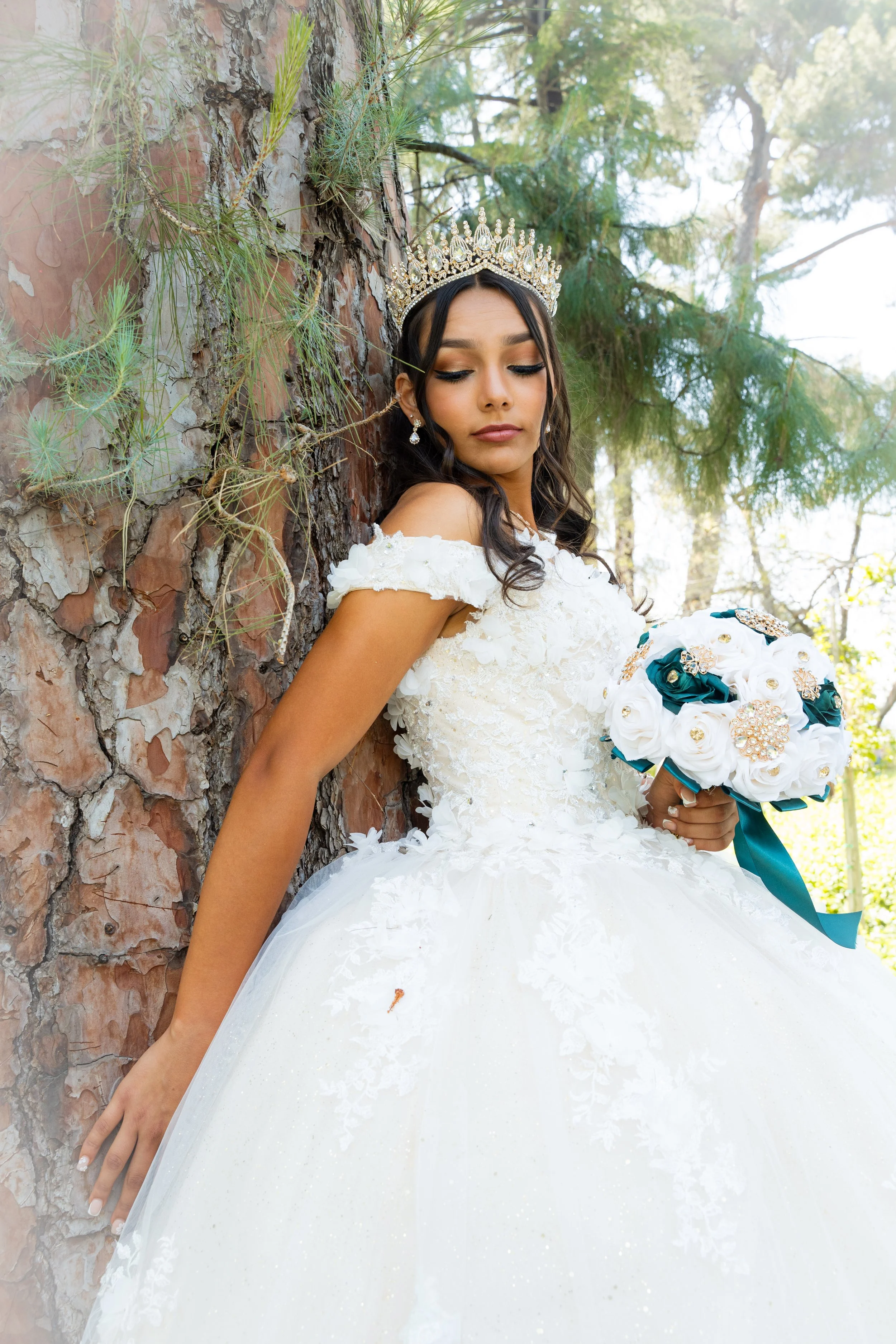 A bride in a white wedding dress, wearing a crown, holding a bouquet, and standing against a tree in a natural setting with green foliage. Redlands California Quinceañera ( Quinceañera photographer)
