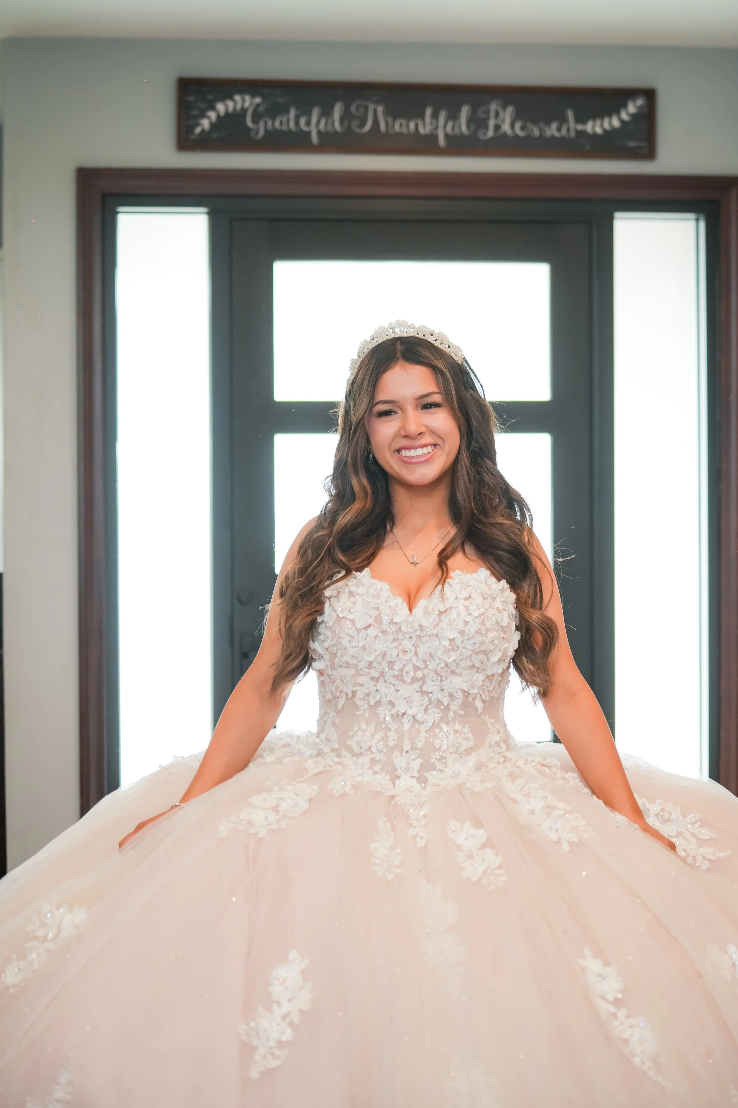 A smiling young woman in a wedding dress, standing indoors in front of a window with a sign above that reads 'Grateful, Thankful, Blessed'. Rialto California Quinceañera ( quinceañera videography)