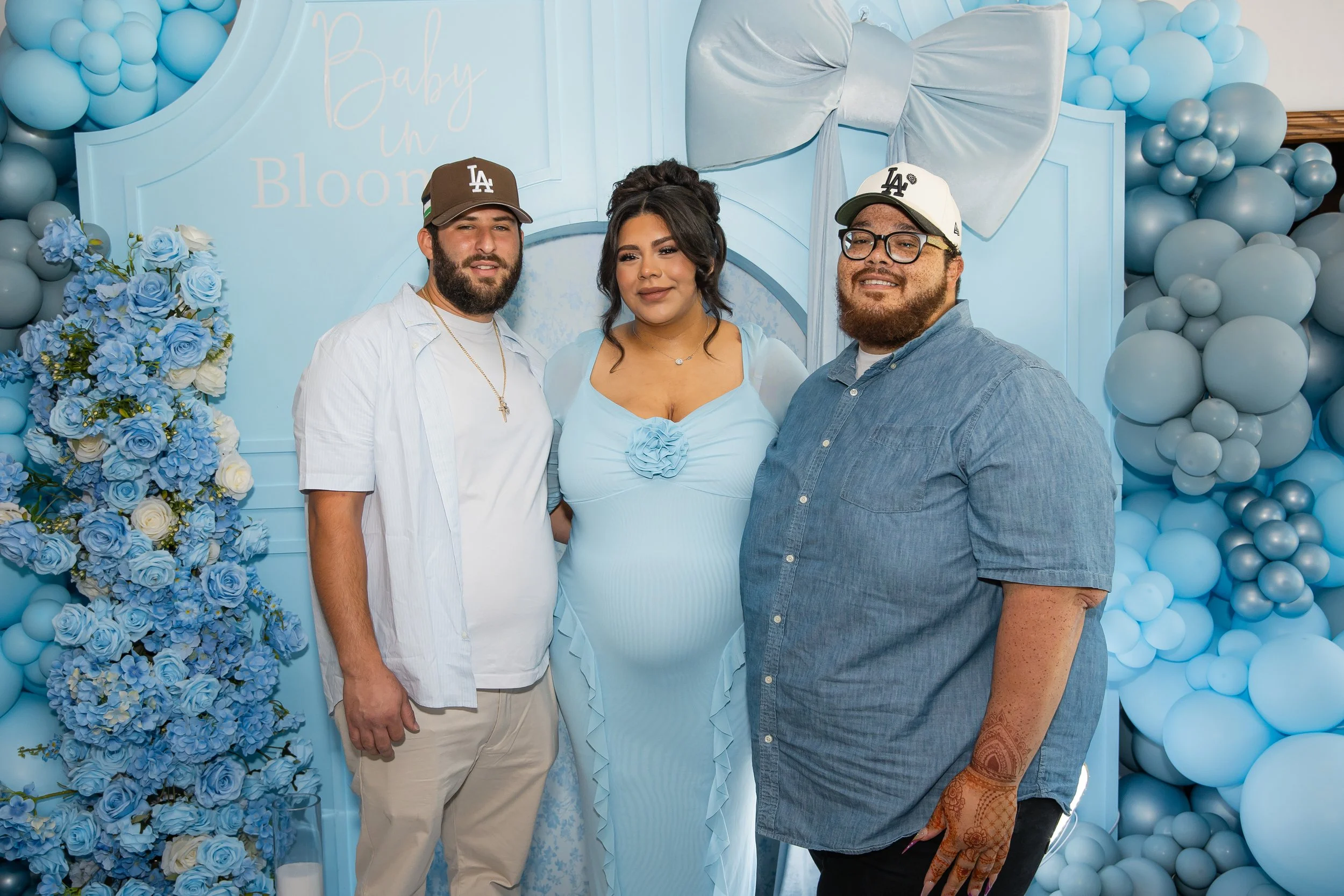 Three people, two men and one pregnant woman, standing together in front of a blue balloon and floral background that says 'Baby in Bloom' at a baby shower. All are smiling, with the woman in a light blue dress, and the men in casual shirts and baseball caps.