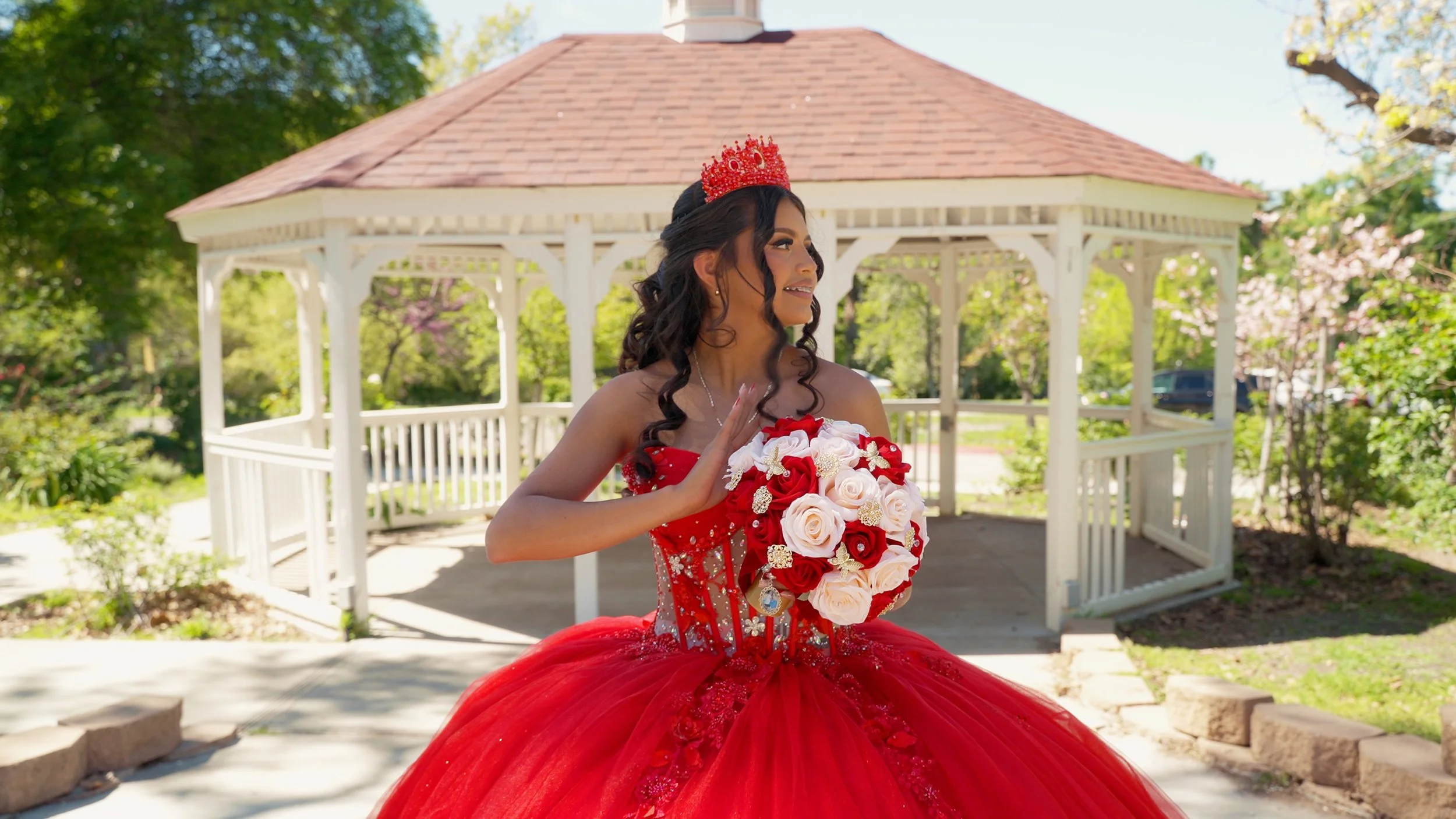 Young woman in a red ball gown with a crown on her head, holding a bouquet of red and white roses, standing outside near a gazebo in a garden. El Monte California Quinceañera ( Quinceañera videographer)