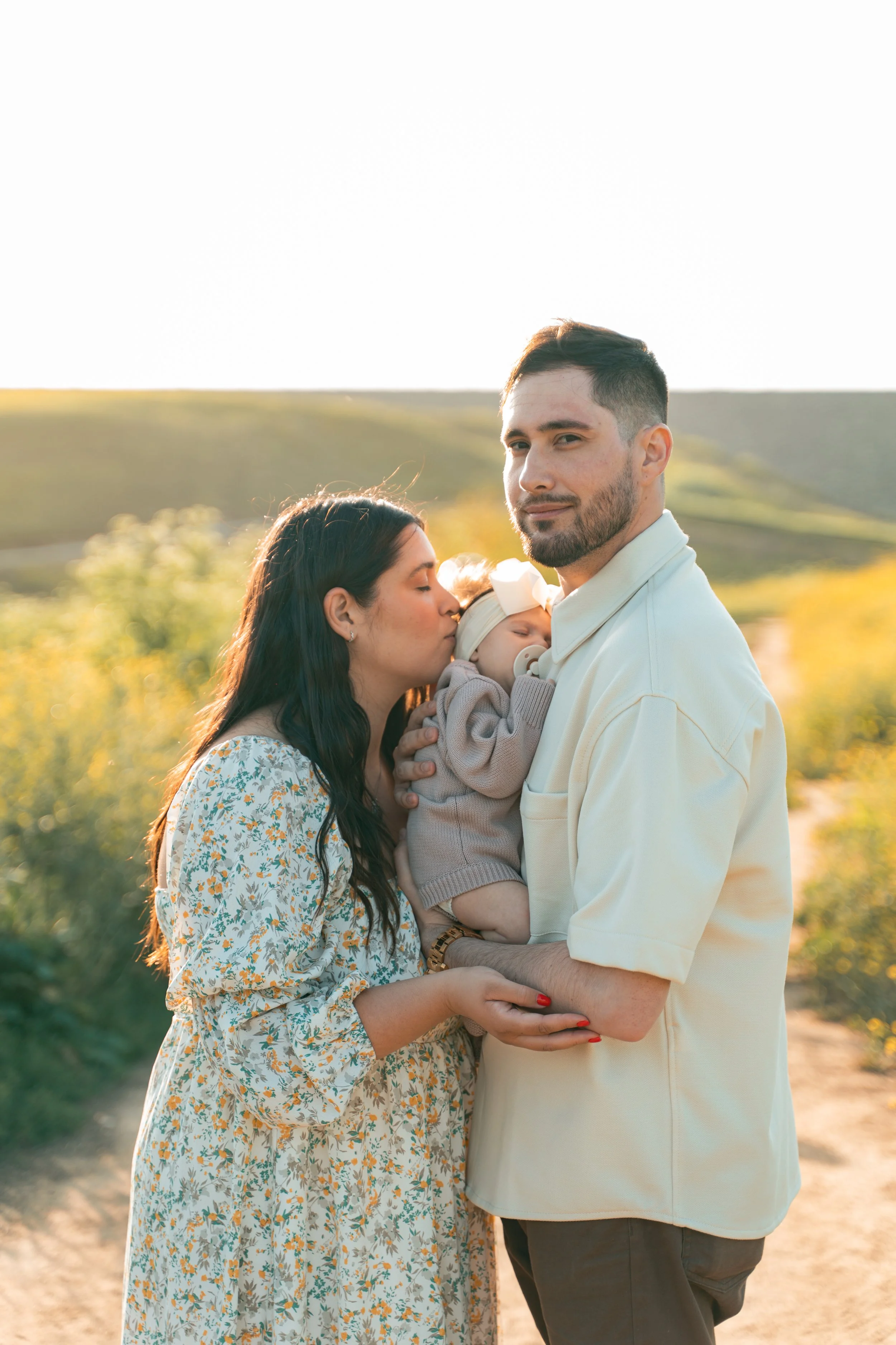 A family of three outdoors in a sunny landscape. The mother is kissing a sleeping baby in the father's arms, with the father looking at the camera. Family portraits Corona California (Local Photographer)