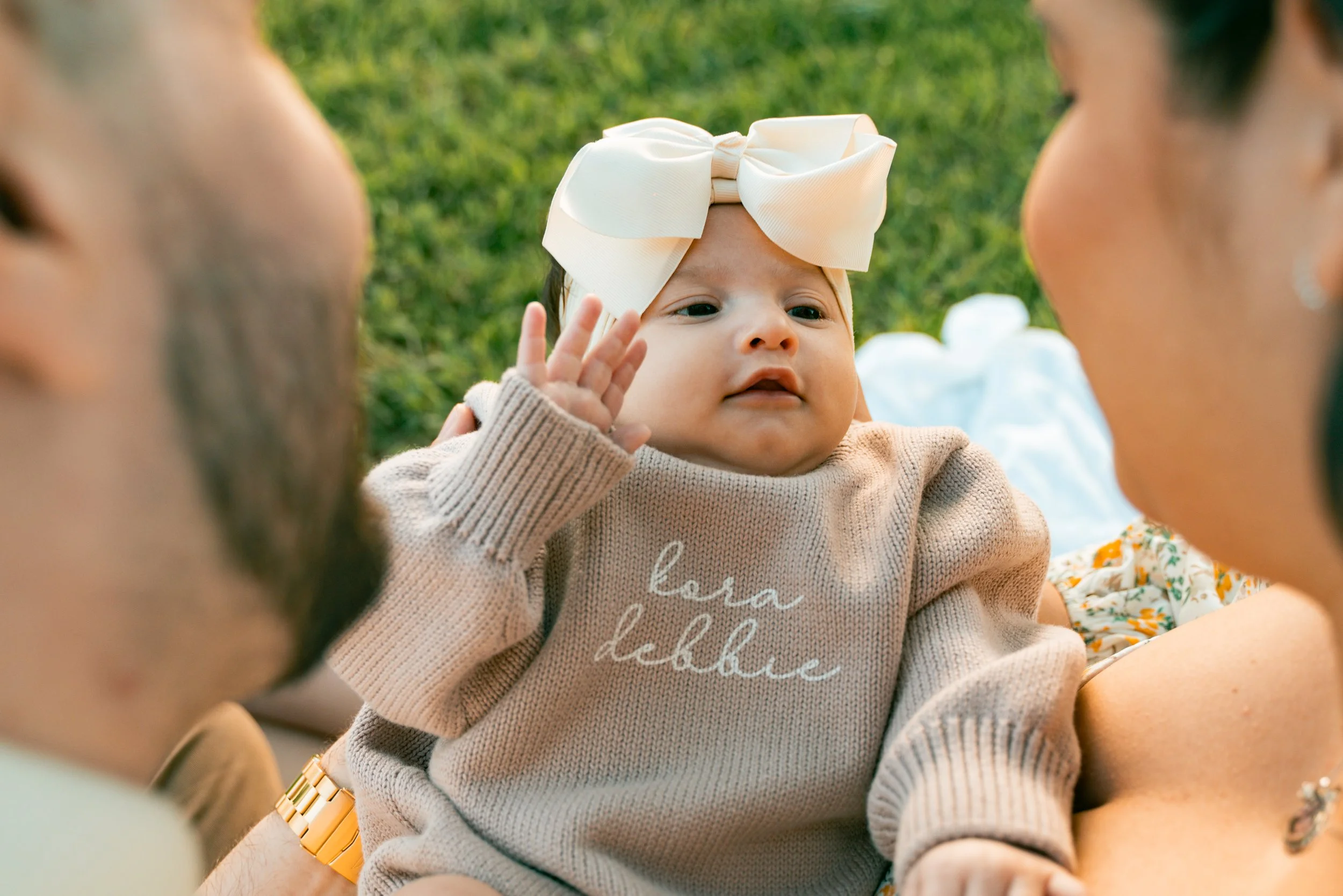 Two adults holding a baby girl with a big beige bow on her head, outside on grass, touching her hand to her face, wearing a beige sweater with the words 'bora debbie' embroidered on it. Family portraits Corona California (Local Photographer)