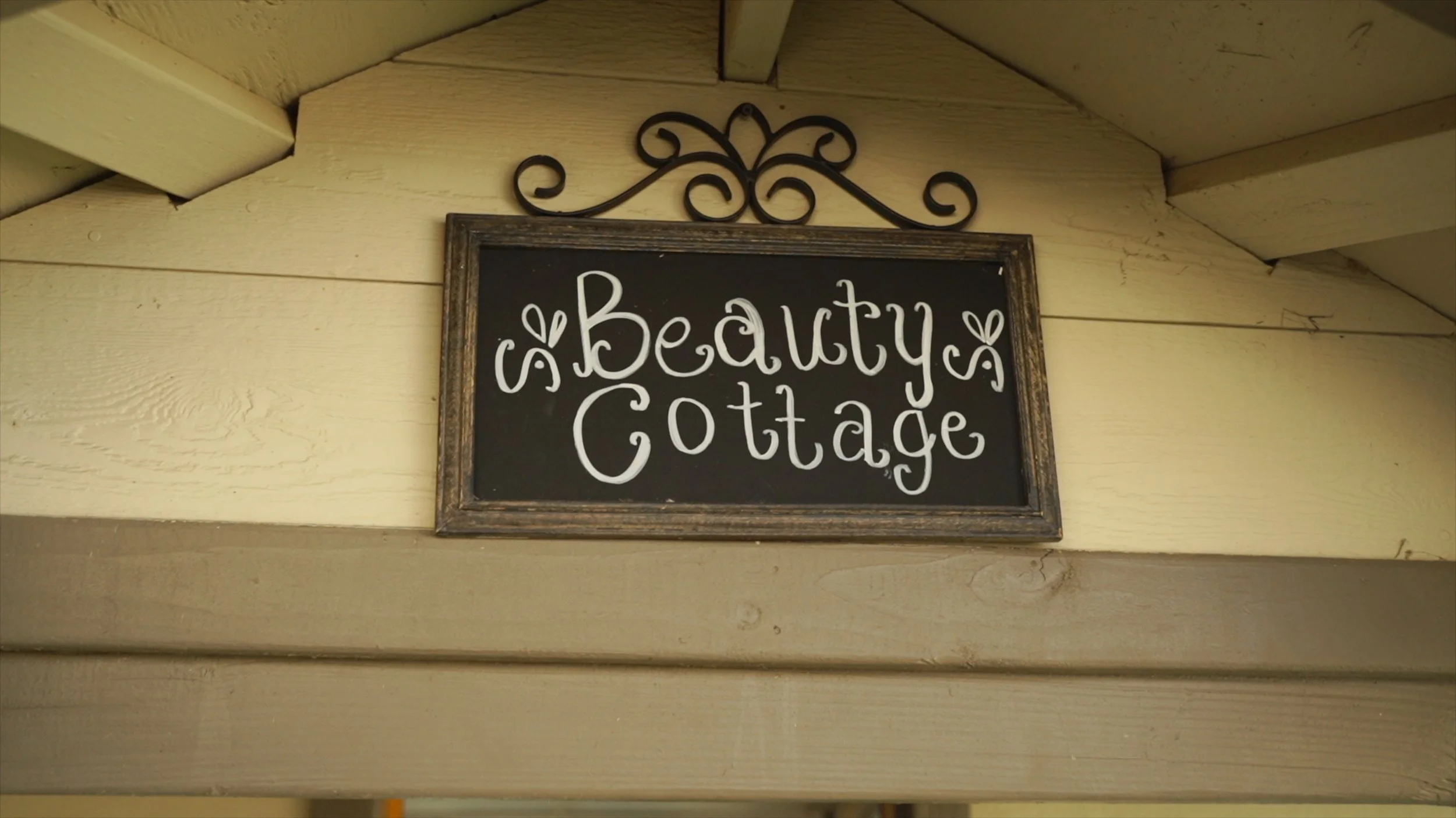 Signboard with the words 'Beauty Cottage' written in decorative white cursive letters on a black background, framed with wood, hanging on a beige wooden wall.