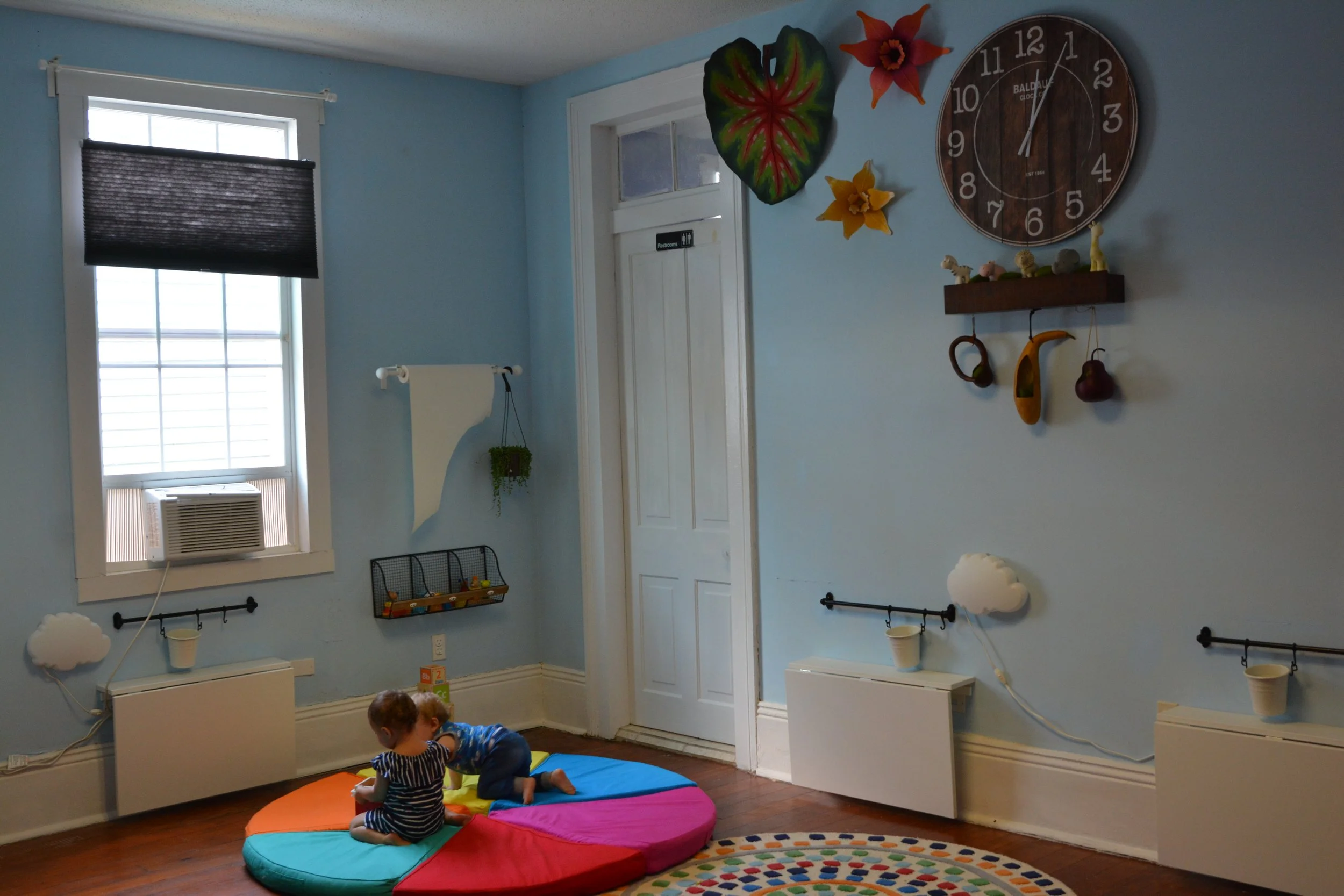 Children playing on a rainbow-colored round cushion in a light blue children's playroom with wall decor, window with black shade, and hardwood floors.