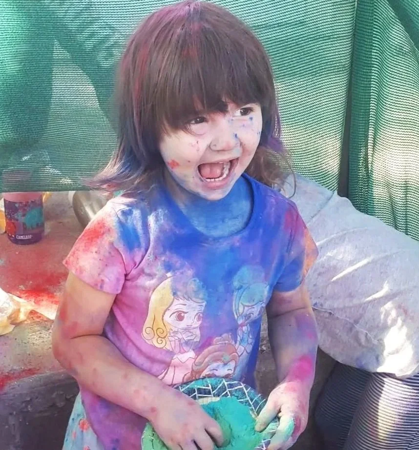 A young girl covered in colored powders, celebrating a festival, with a big smile and messy face.