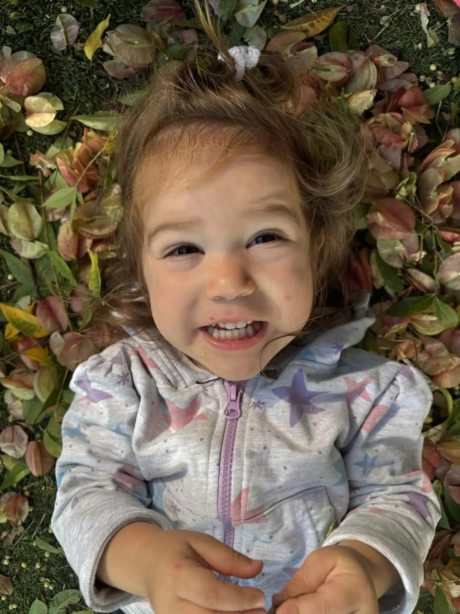 A young girl with curly hair and a butterfly hair clip lying on grass and fallen leaves, smiling and looking up at the camera.
