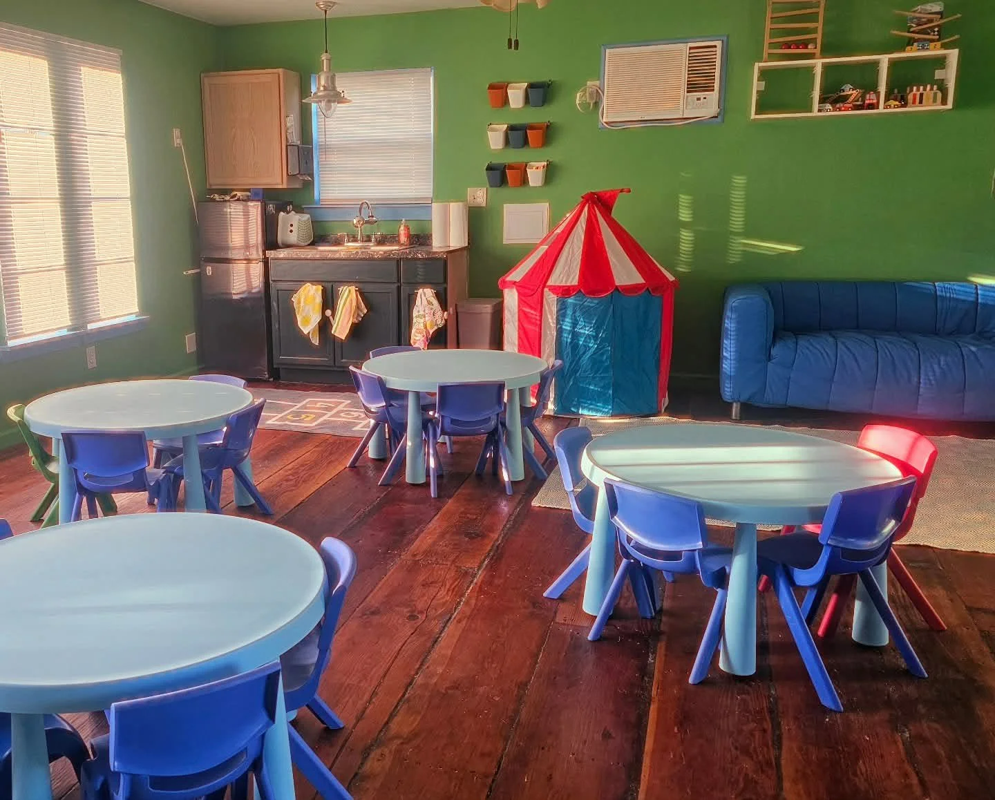 Indoor play area with small blue tables and chairs, a red and white play tent, a blue couch, a kitchenette with a sink, and colorful wall decorations.