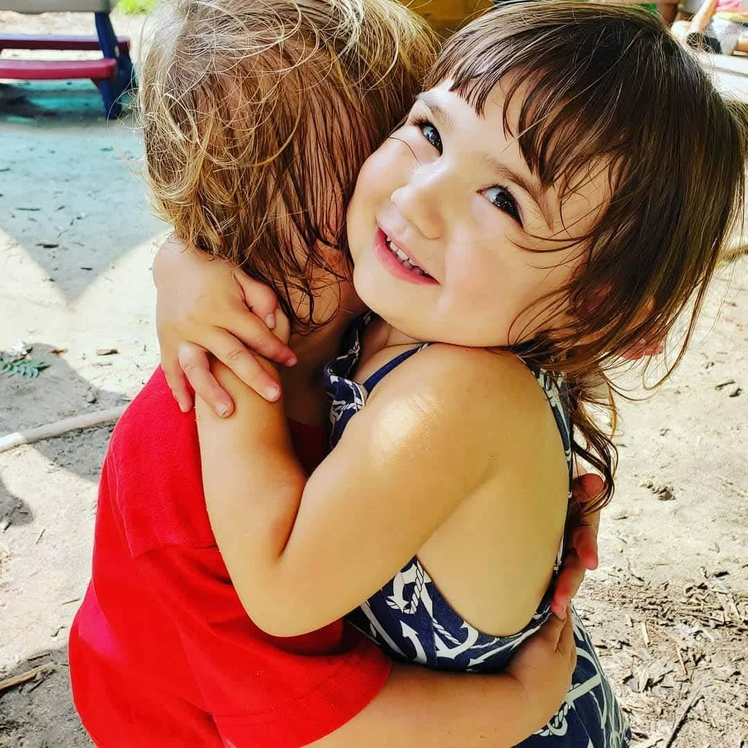 Two young children hugging and smiling on a sandy outdoor playground.
