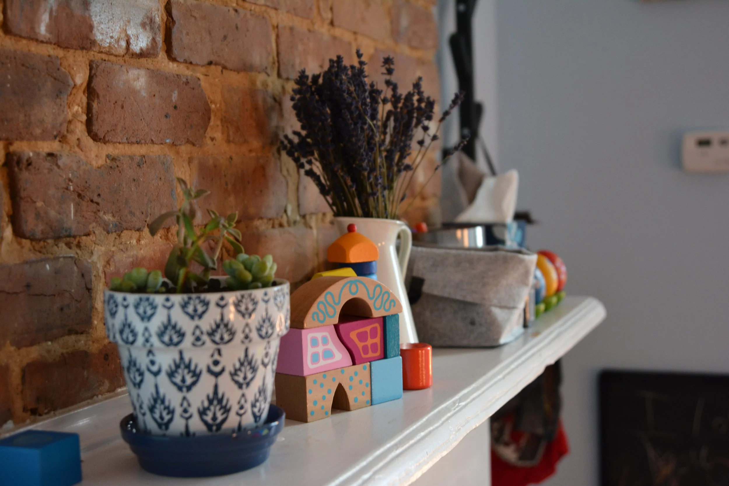 A shelf against a brick wall displaying a small potted plant with succulent leaves in a blue and white patterned pot, a colorful toy house, and a bouquet of dried purple flowers in a white mug.