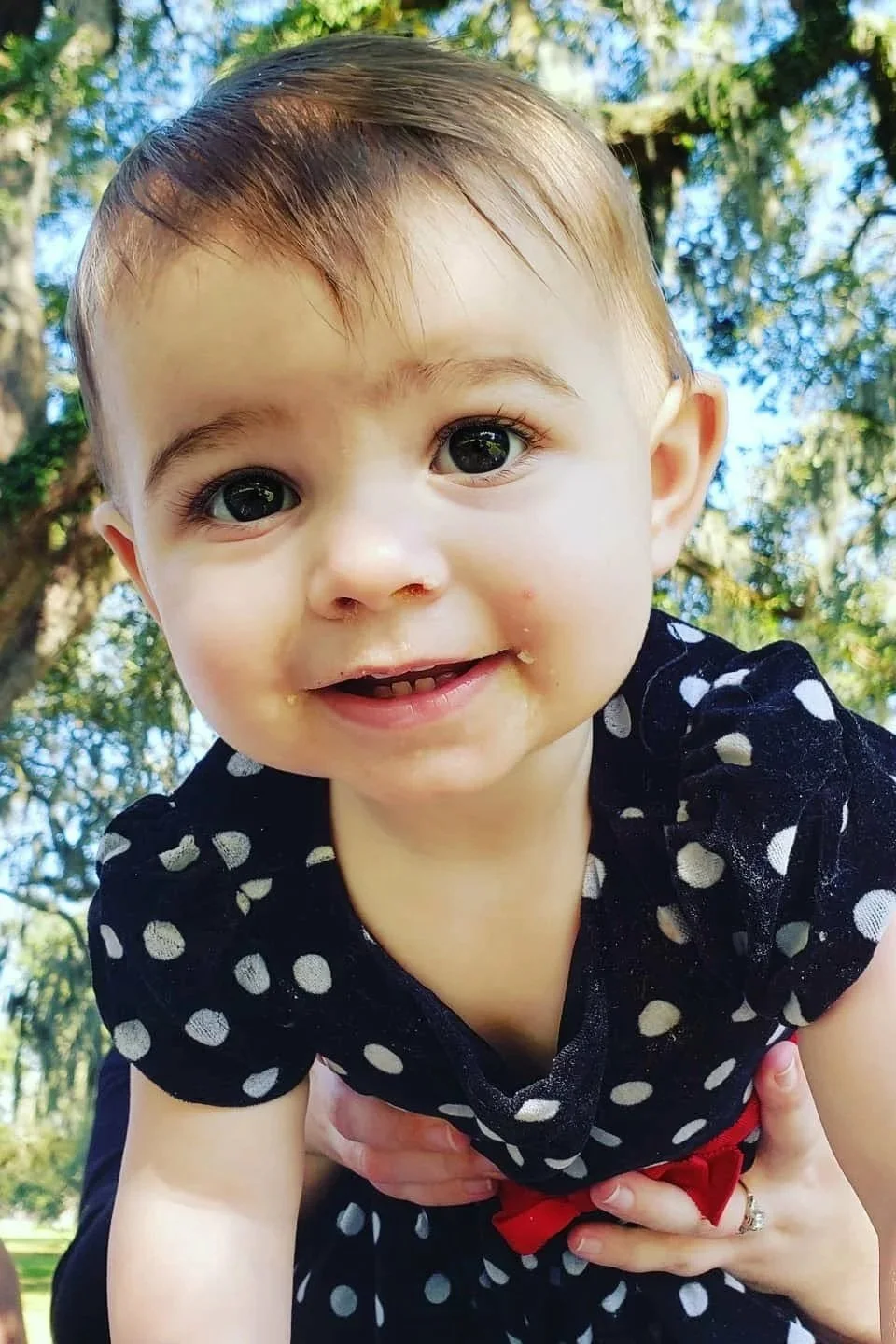 Close-up of a smiling toddler girl with big eyes, brown hair, wearing a black dress with white polka dots, outdoors with trees in the background.