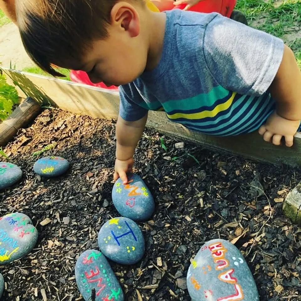 A young boy in a striped shirt is painting colorful designs on rocks placed in soil within a garden bed.