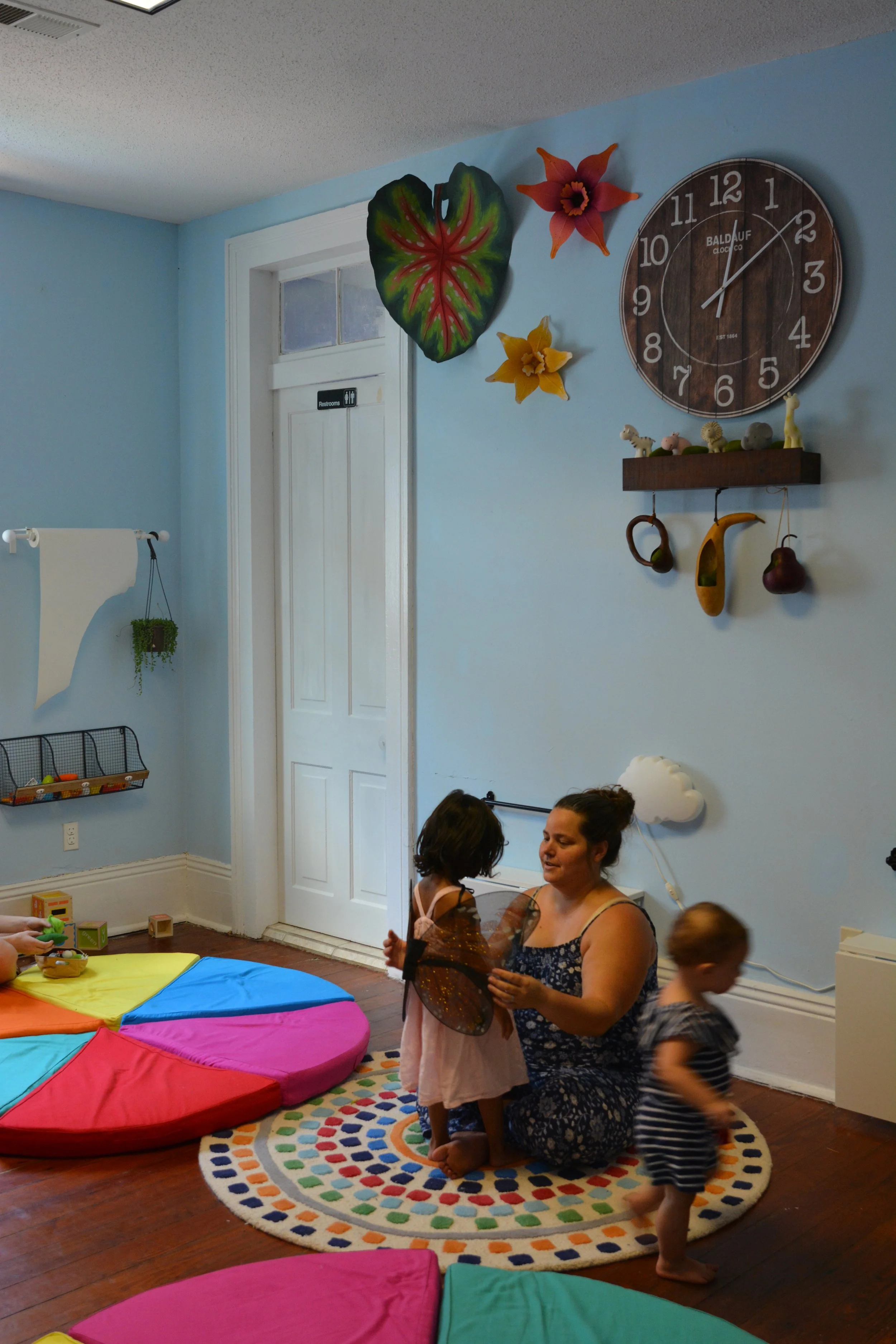 Children and adults inside a room with light blue walls, colorful cushions on the floor, a rainbow-colored circular rug, and wall decorations including a large clock, hanging figurines, and paper flowers. A woman is holding a child with butterfly win