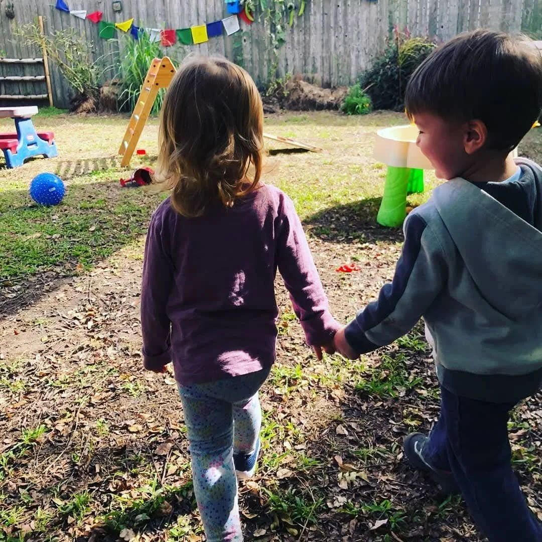 Two young children, a girl and a boy, holding hands and walking in a backyard play area with toys and a wooden fence in the background.