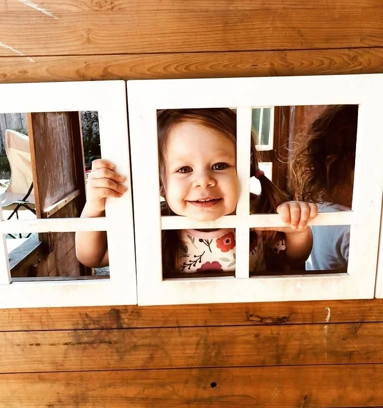 A smiling young girl with brown hair looking through a small window in a wooden playhouse, holding the window frame with her hands.
